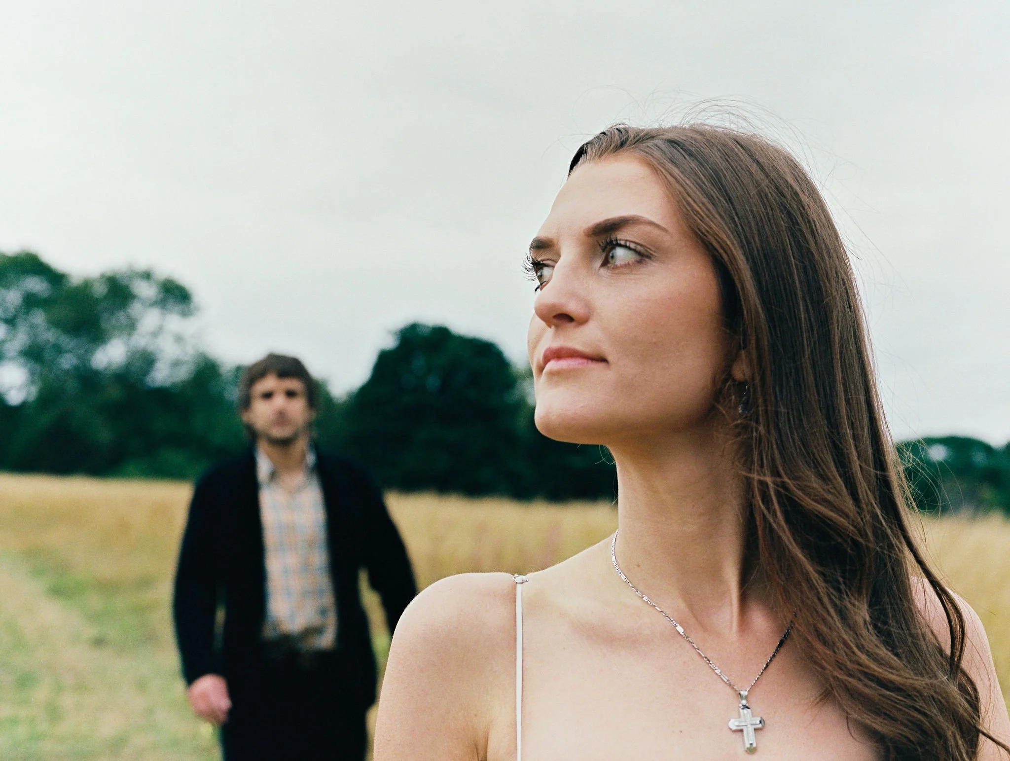 A woman with long brown hair wearing a necklace with a cross pendant looking to the side in a field with trees in the background, and a man in a dark suit and checkered shirt in the distance.