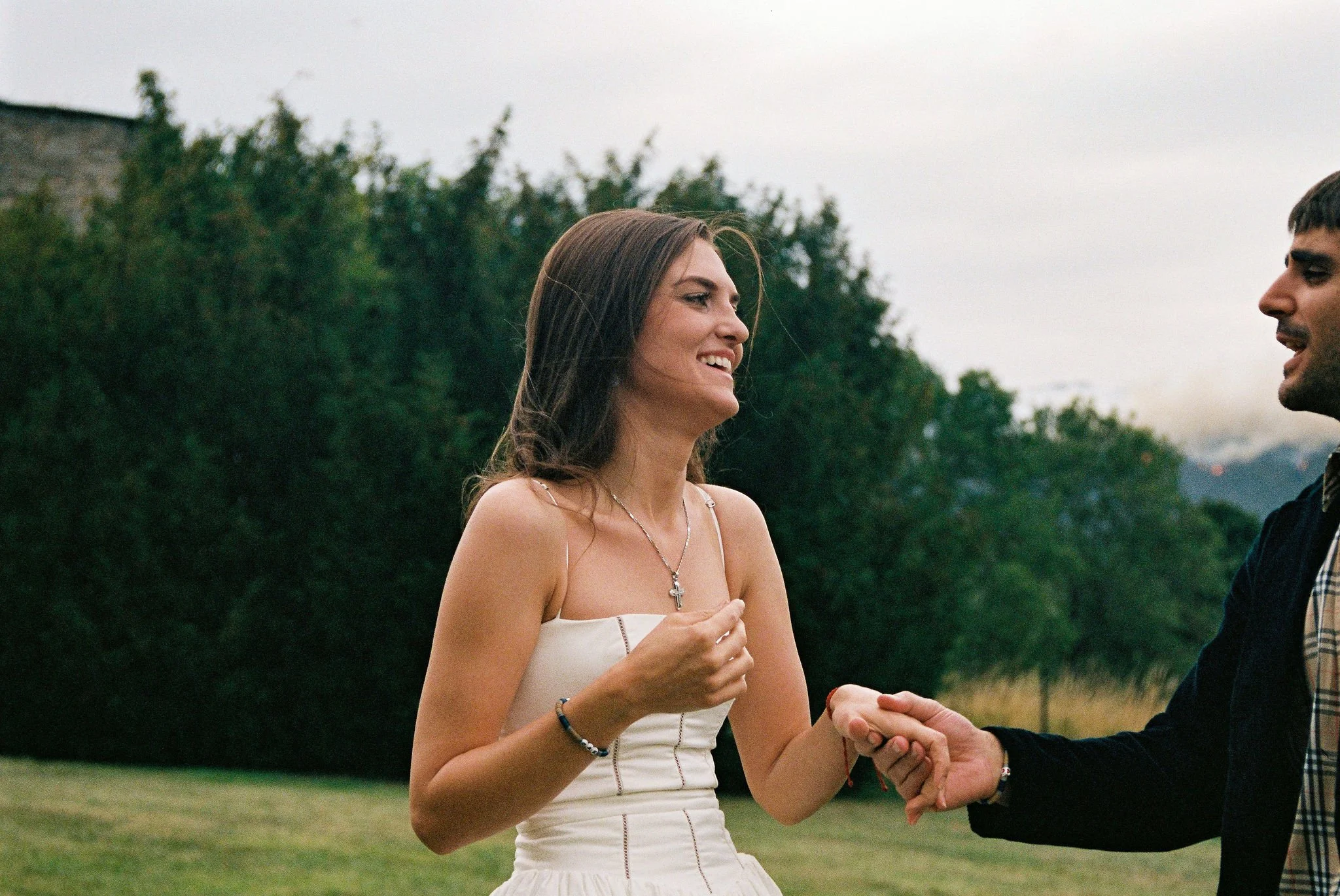 A woman in a white dress with a cross necklace smiling and holding hands with a man outdoors in a grassy area with trees in the background.