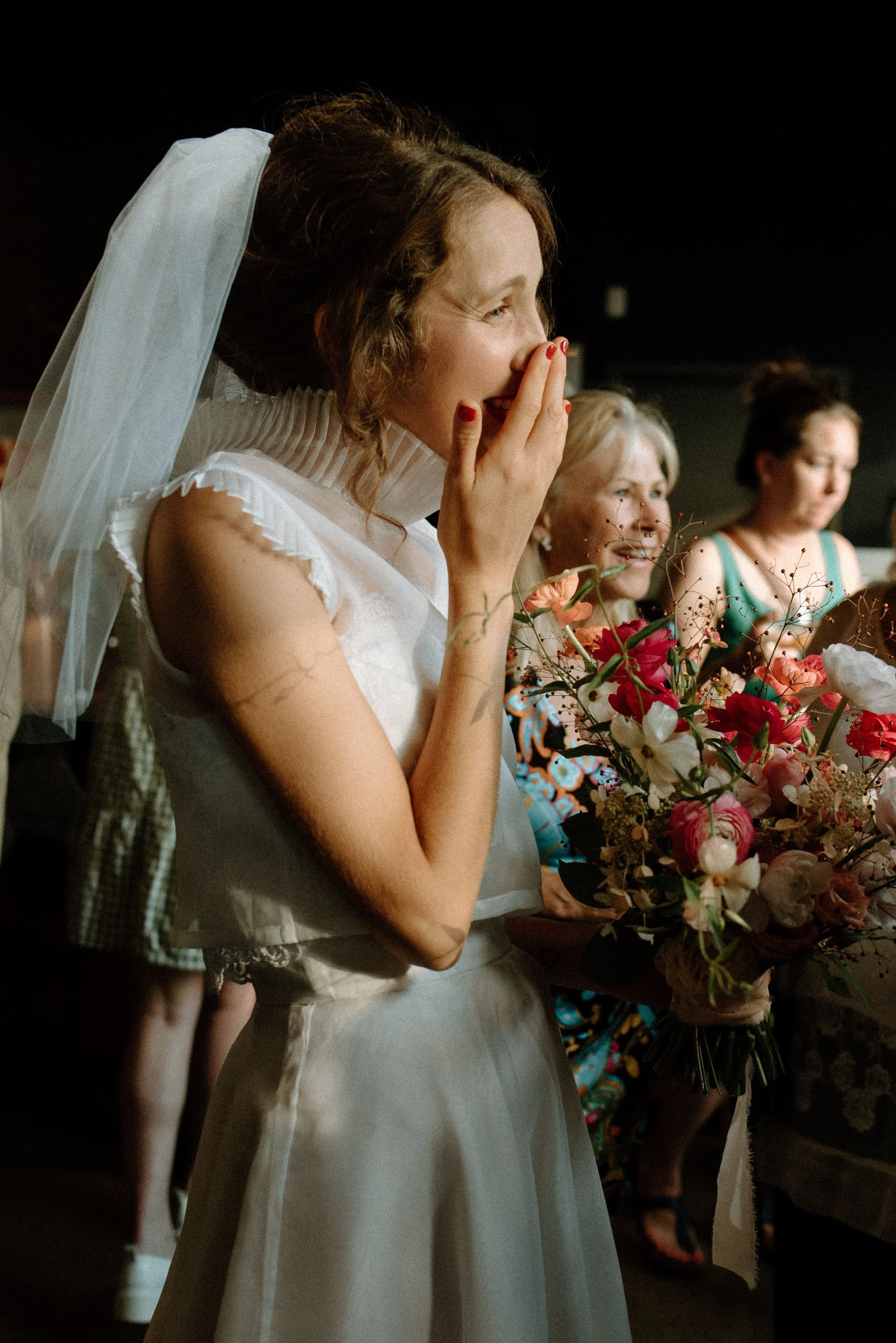 A woman in a white dress and veil, holding a bouquet of flowers, is emotional and surprised, with her hand covering her mouth, at a wedding reception.