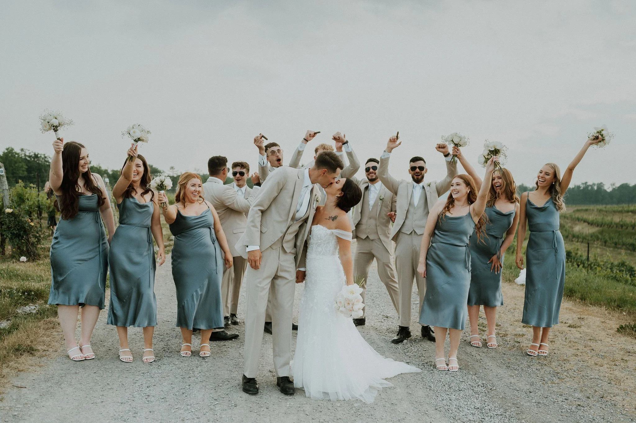 A wedding party with the bride and groom kissing in the center, surrounded by bridesmaids and groomsmen celebrating outdoors on a gravel path.