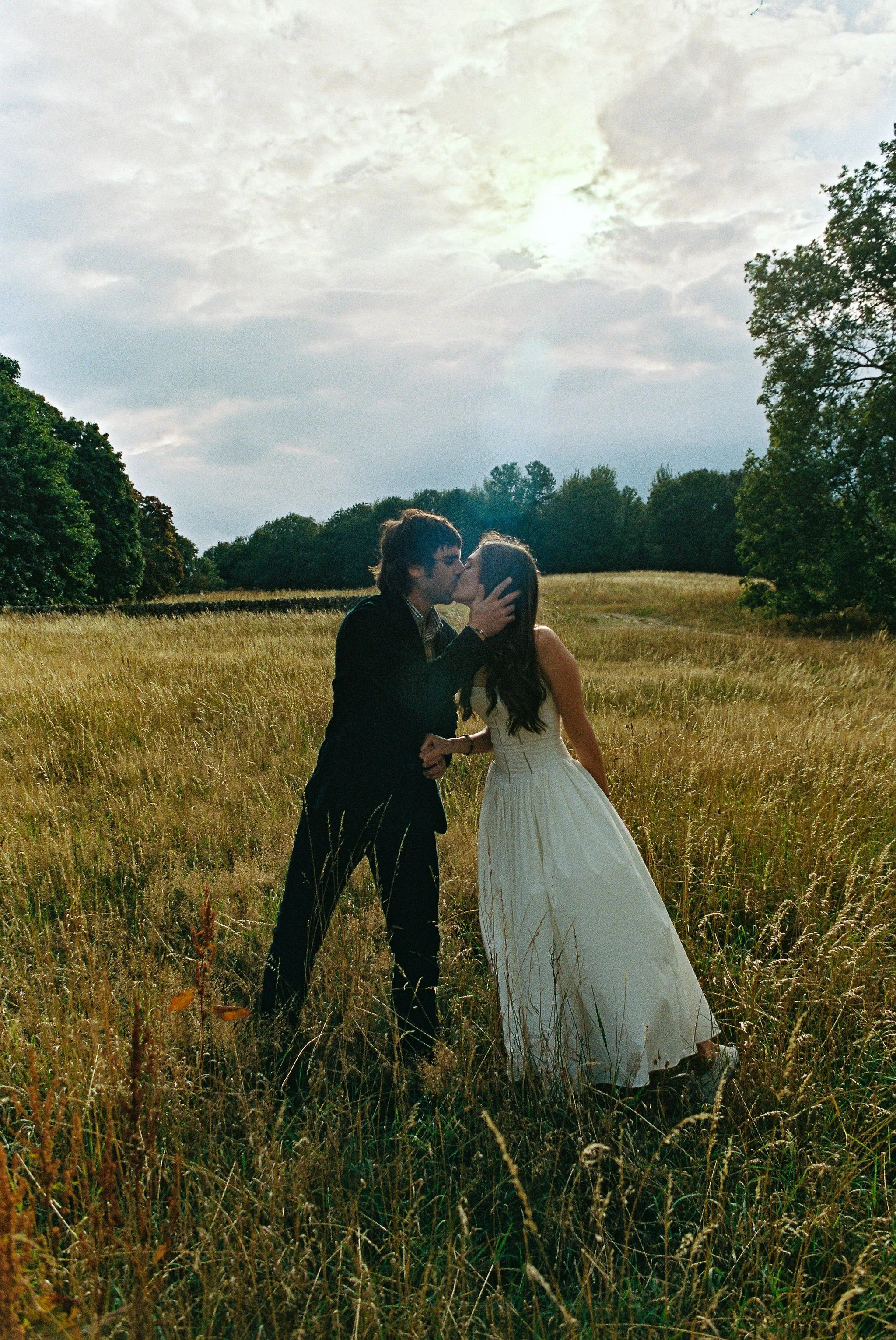 A couple in formal attire sharing a kiss in a grassy field with trees in the background and sky overhead.