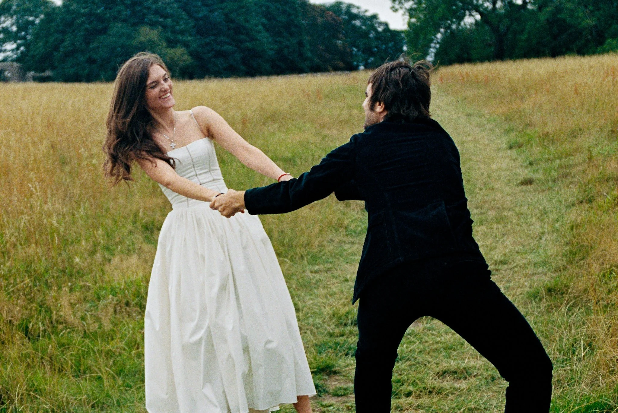 A woman in a white dress and a man in a black jacket are holding hands and playing in a grassy field.