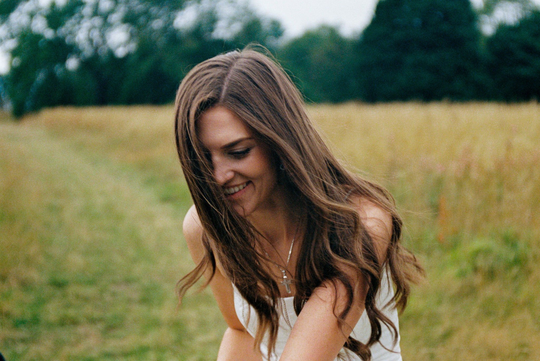 A woman with long brown hair smiling outdoors in a field with trees in the background.