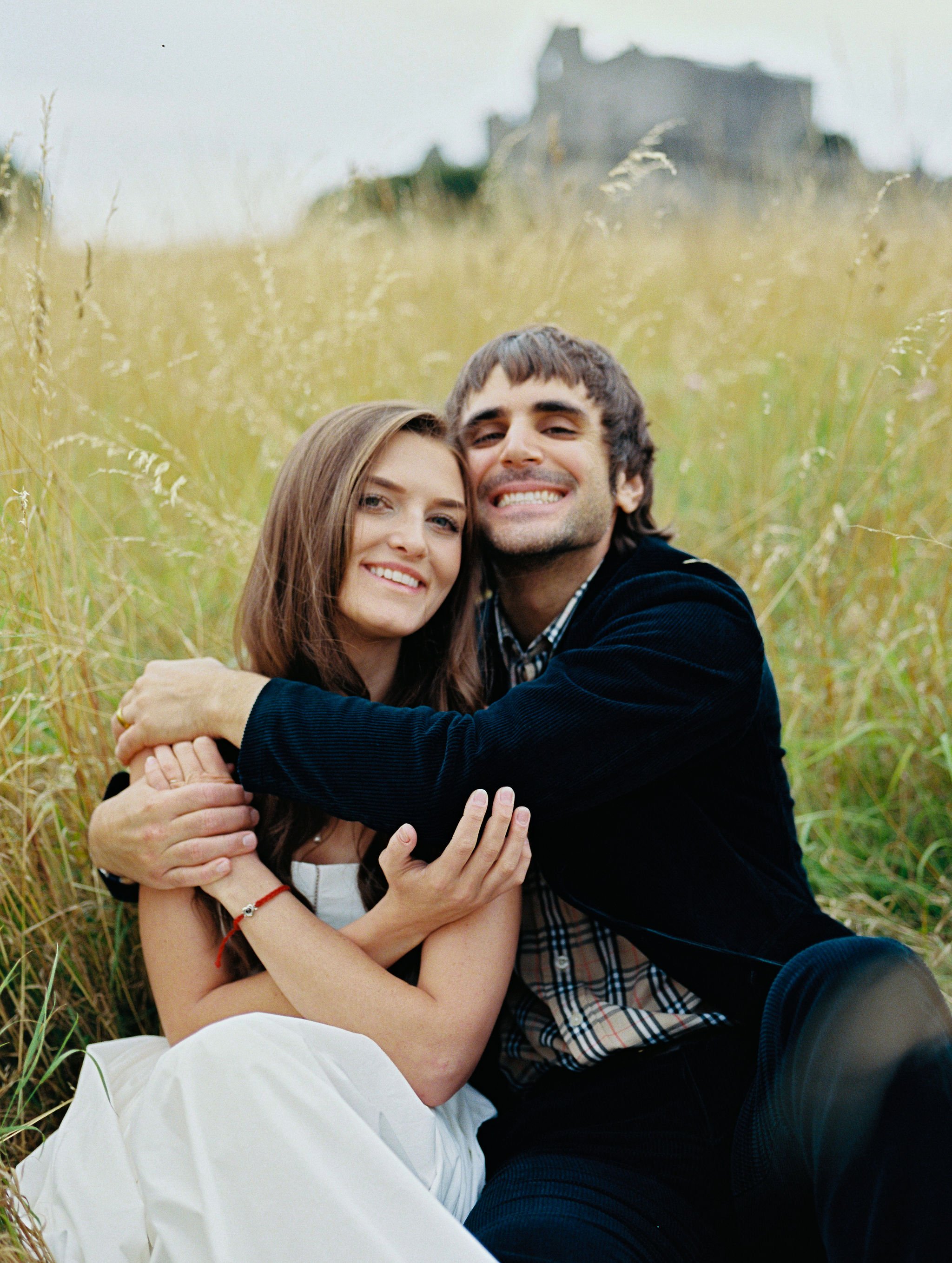 A smiling couple sitting together in a grassy field with tall yellow grass, embracing each other, with a blurred building or structure in the background.