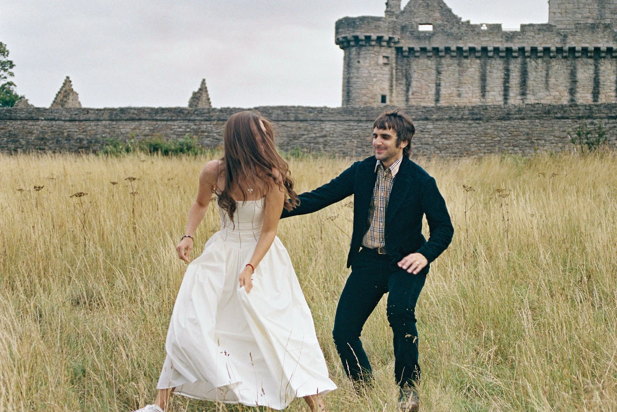 A couple dancing in a grassy field near a stone castle, with the woman in a white dress and the man in dark clothing.