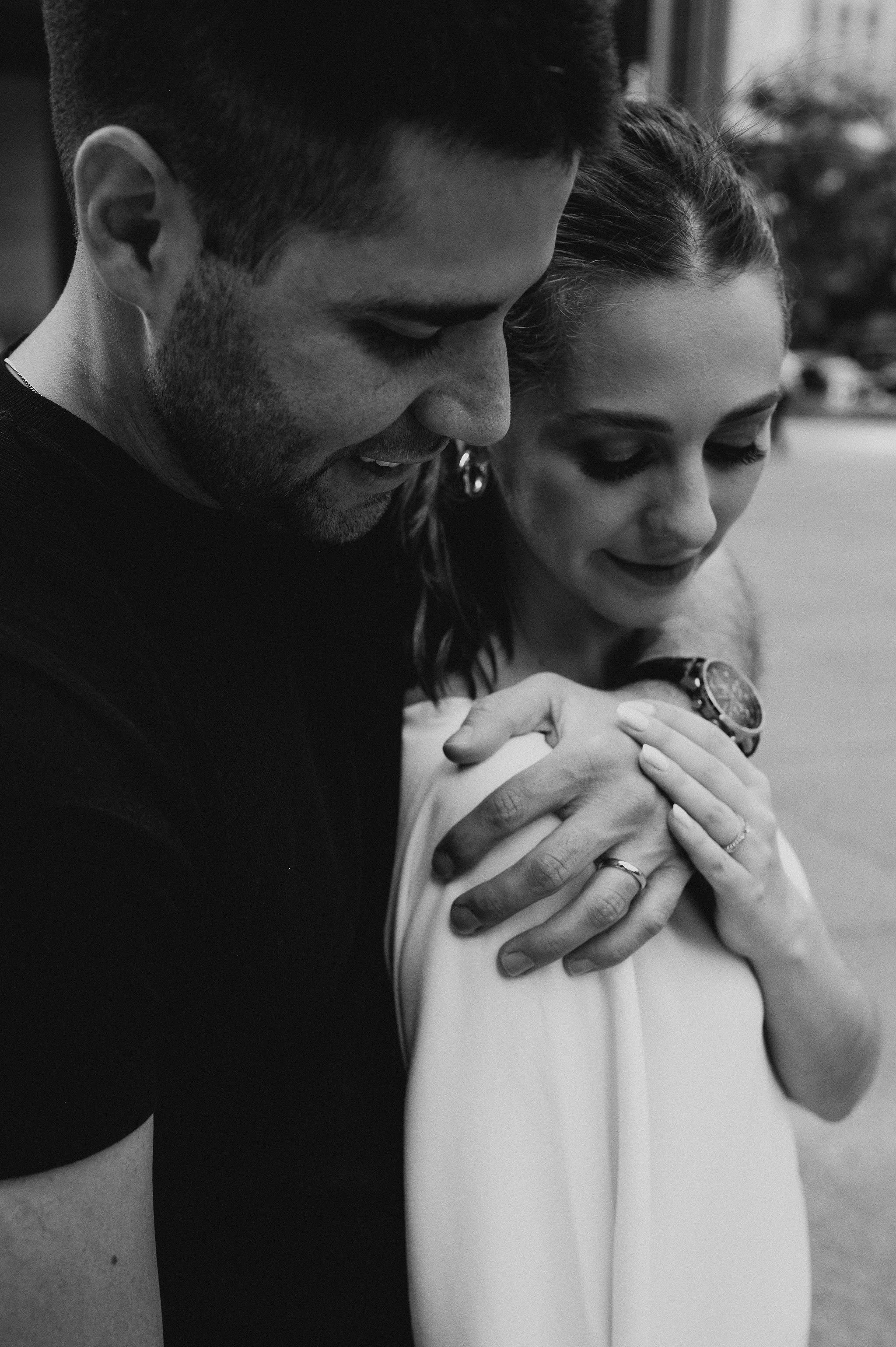 A black and white photo of a man and woman embracing each other, with the man smiling and the woman closing her eyes, both wearing wedding rings.
