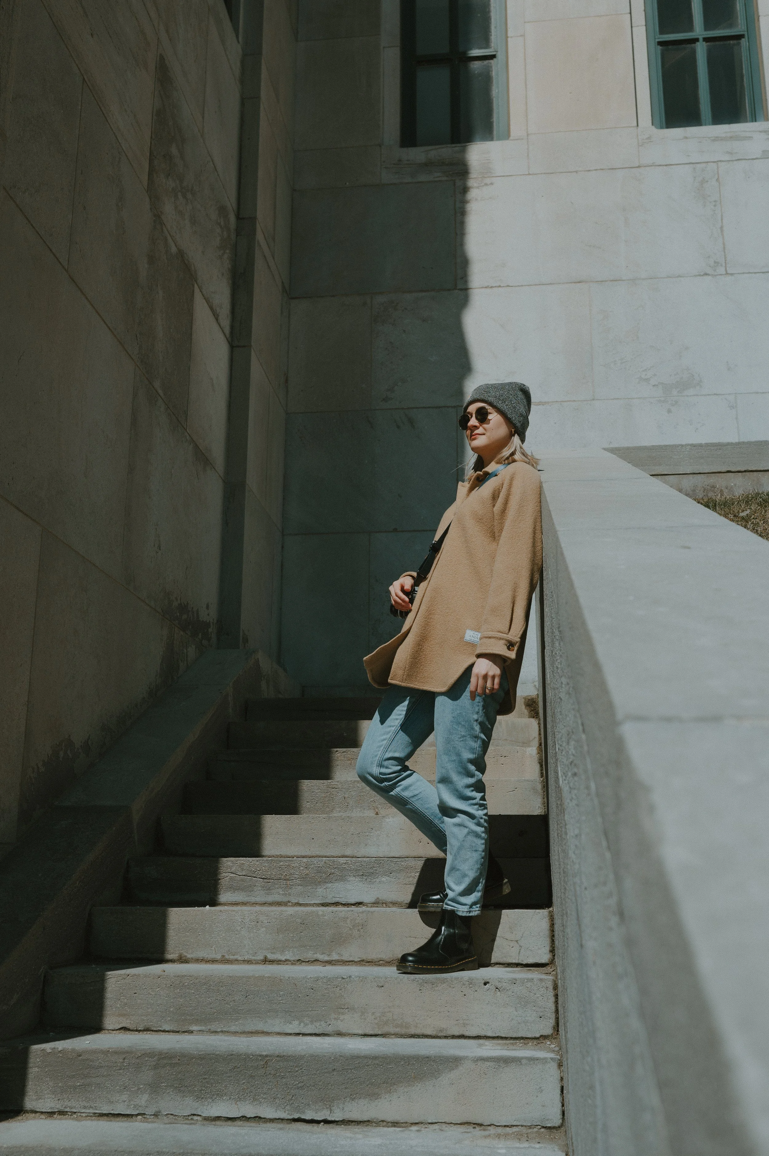 A young woman wearing a gray beanie, sunglasses, a tan coat, blue jeans, and black boots standing on outdoor concrete stairs beside a concrete wall, with sunlight casting a shadow on the wall.