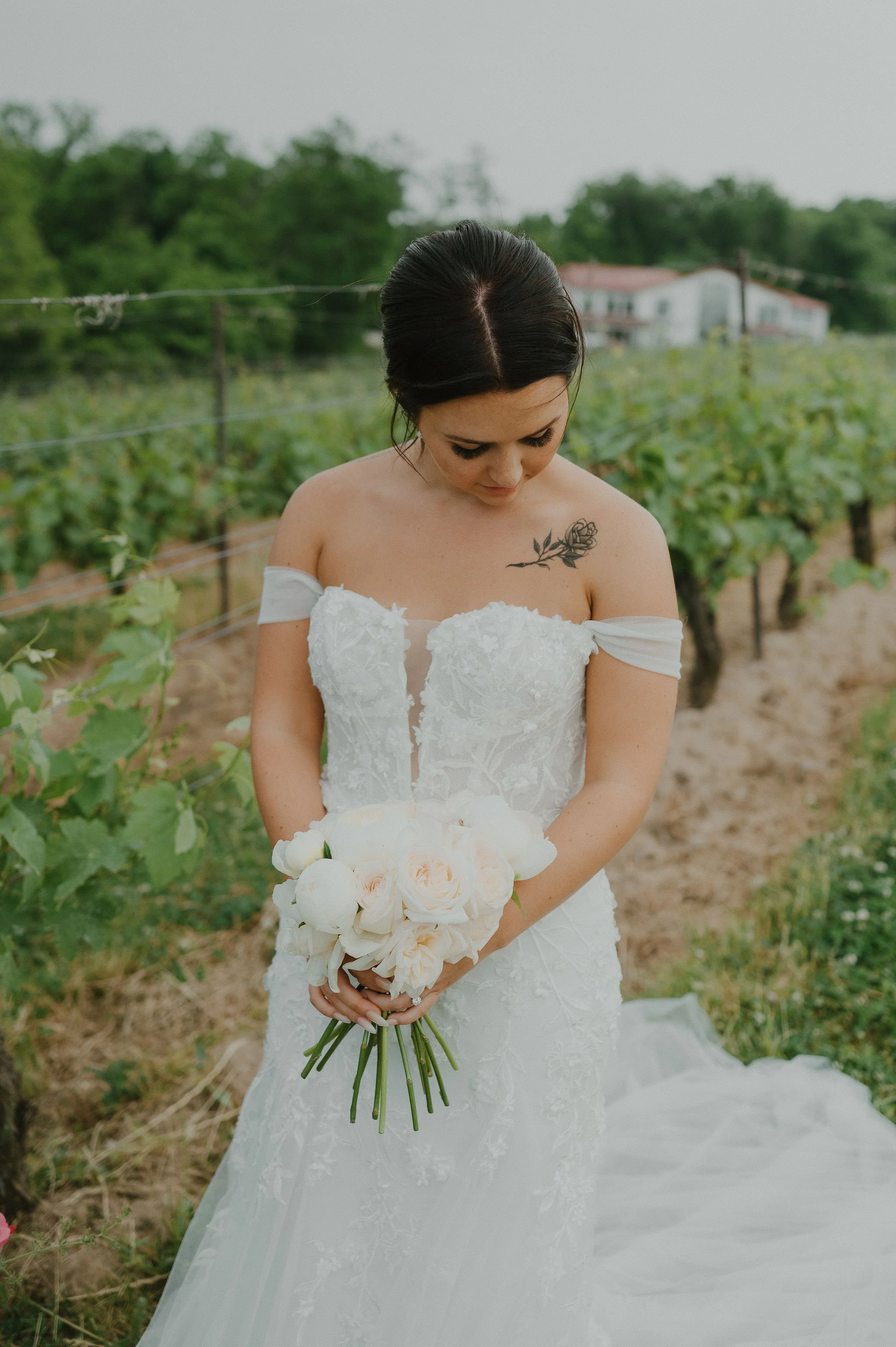 A bride in a white lace wedding gown holding a bouquet of white roses and peonies, standing outdoors in a vineyard with green foliage and a house in the background.