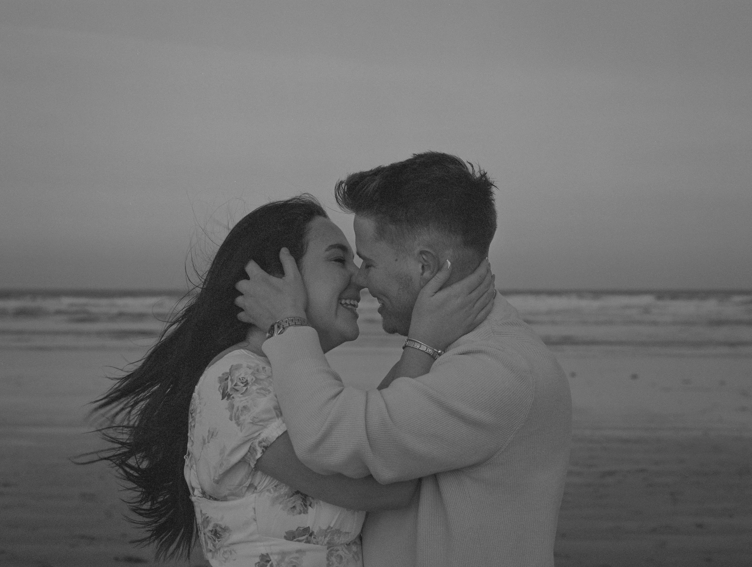 A black and white photo of a happy couple embracing on the beach, smiling and touching foreheads.