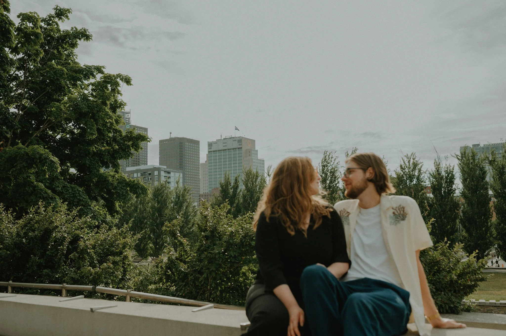 A couple with light skin sitting on a ledge in a park, gazing into each other's eyes with a city skyline in the background, trees, and overcast sky.