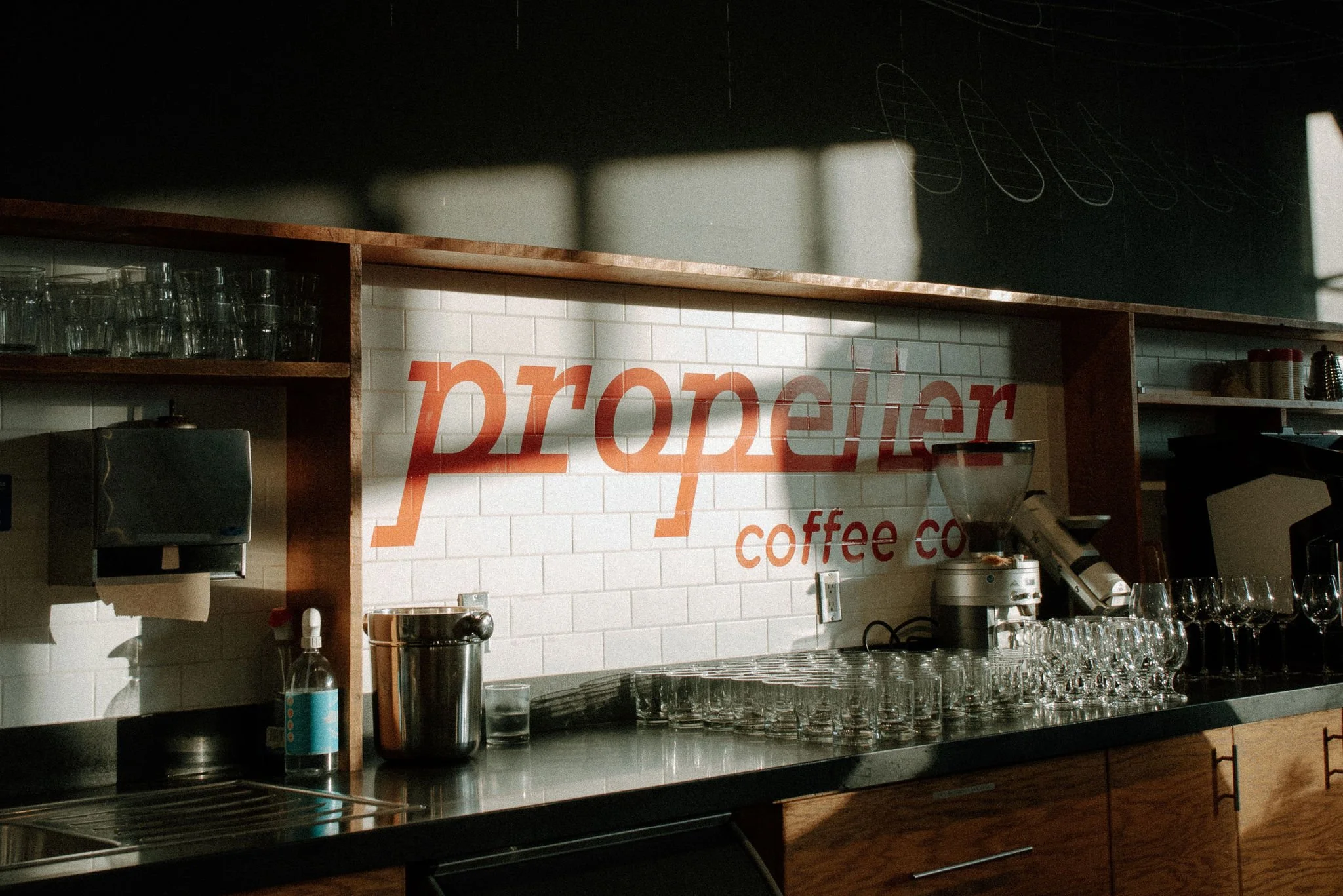 Inside a coffee shop with a counter displaying glasses and coffee equipment, a white tiled wall with red lettering spelling 'propeller' and 'coffee co.'