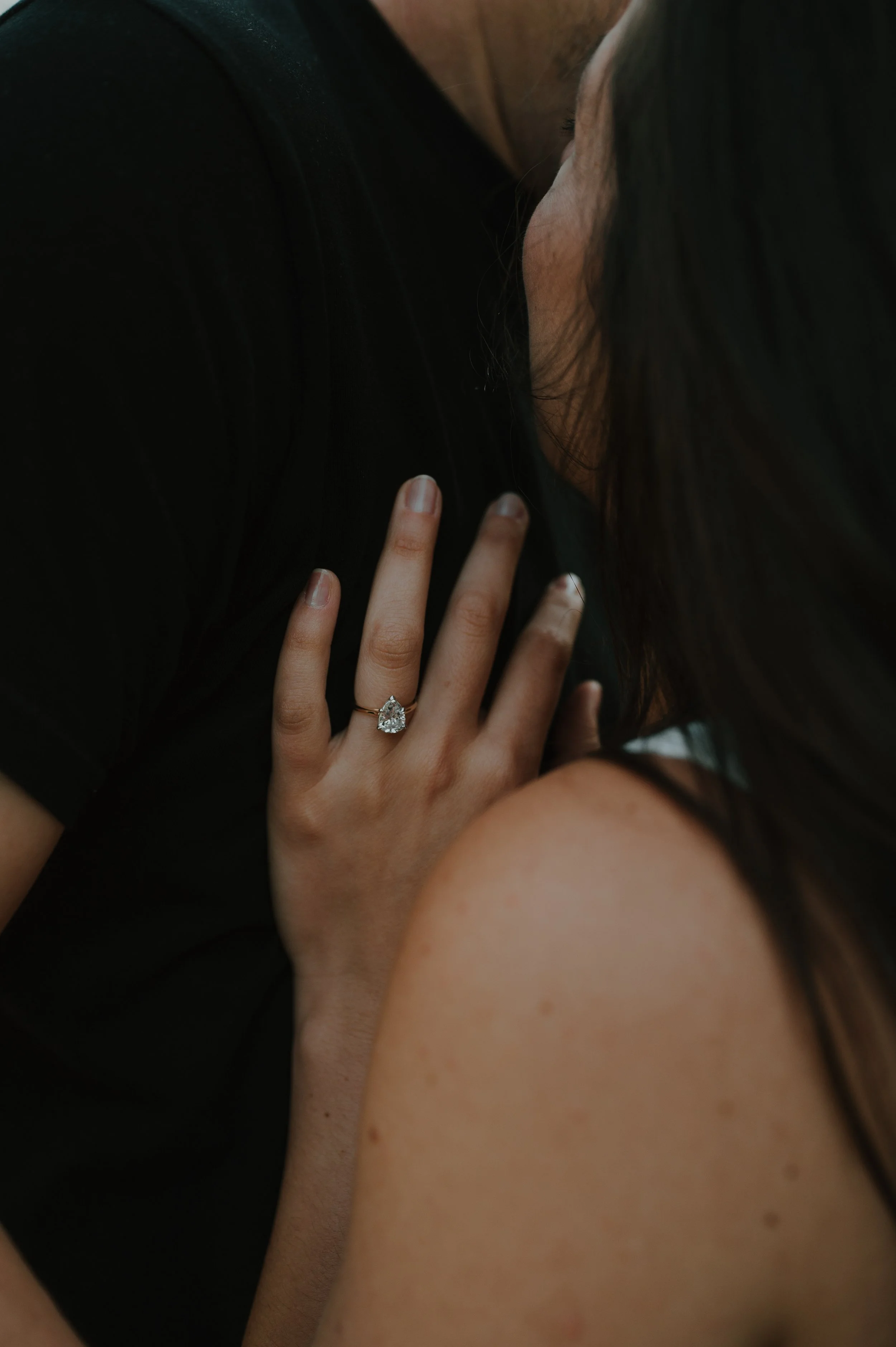 A woman shows off a large, sparkly engagement ring on her finger. Her hand rests on a man's shoulder, and she has long dark hair and lightly tanned skin.
