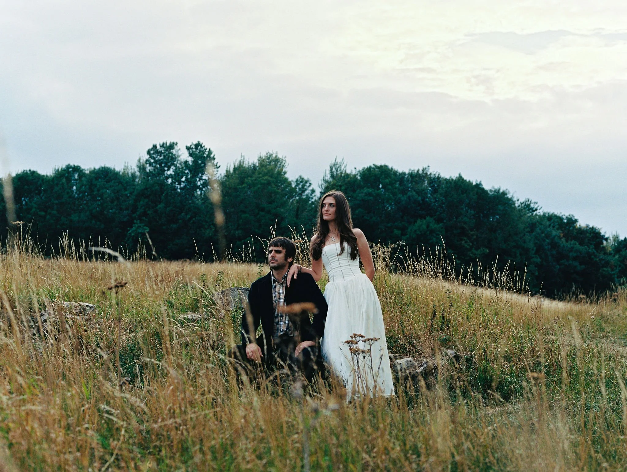 A woman in a white dress standing next to a man in a dark jacket and plaid shirt seated in a grassy field with trees in the background.