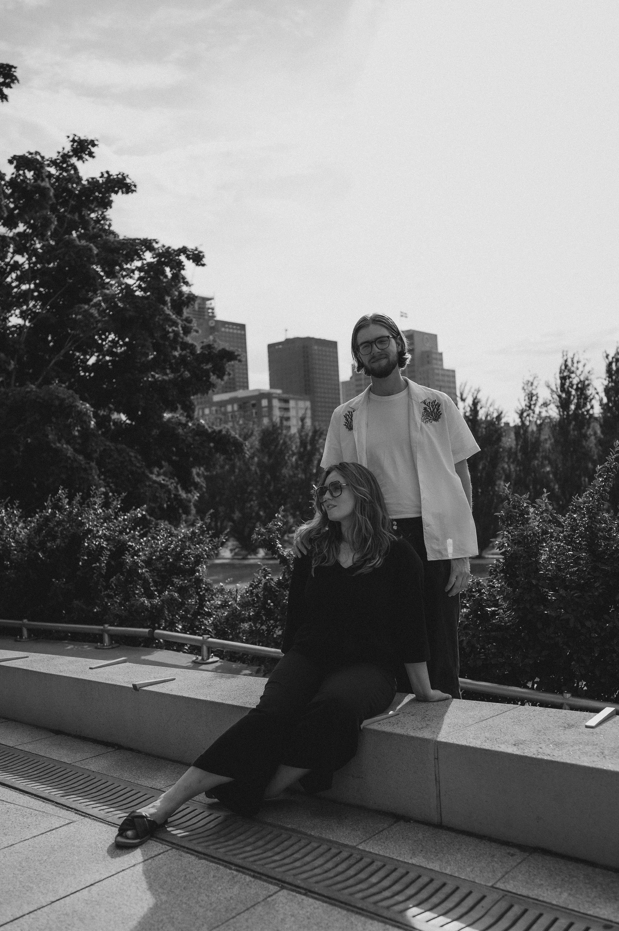 A black and white photo of a man and woman outdoors with city buildings in the background. The woman is seated on a ledge, wearing glasses and dark clothing, while the man stands behind her, wearing glasses and a light shirt with embroidery.