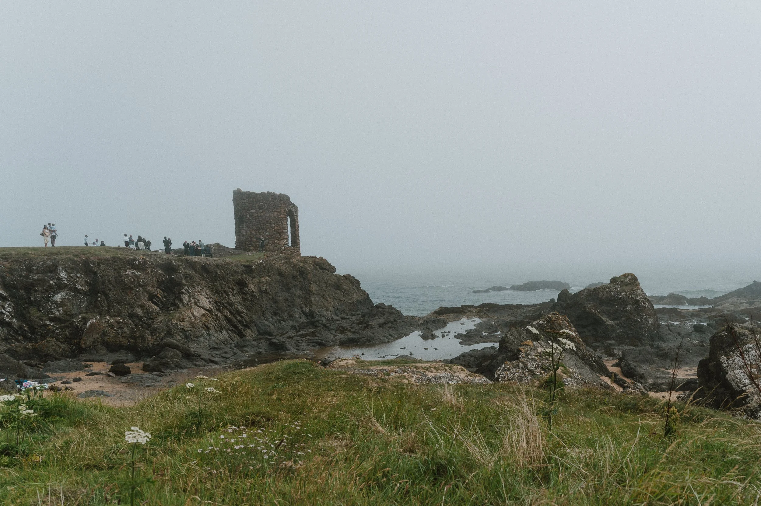 Coastal landscape with rocky shoreline, grassy foreground with wildflowers, and a stone tower with an arched window on a cliff, foggy sky, and people in the distance exploring the area.