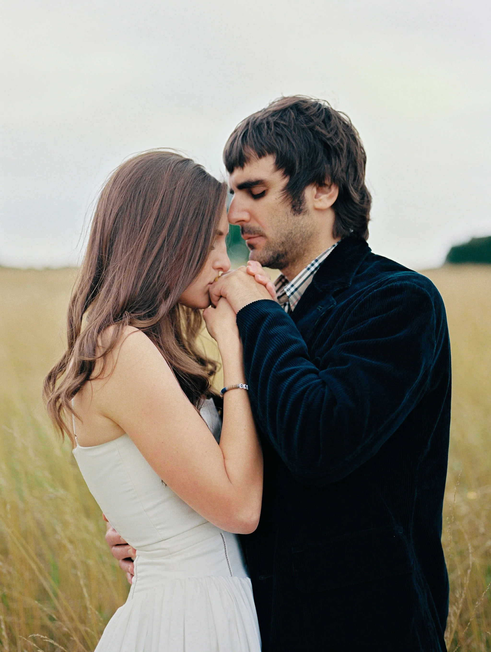 A young woman and man are embracing and holding hands in a field of tall grass on a cloudy day, with their foreheads touching and eyes closed.