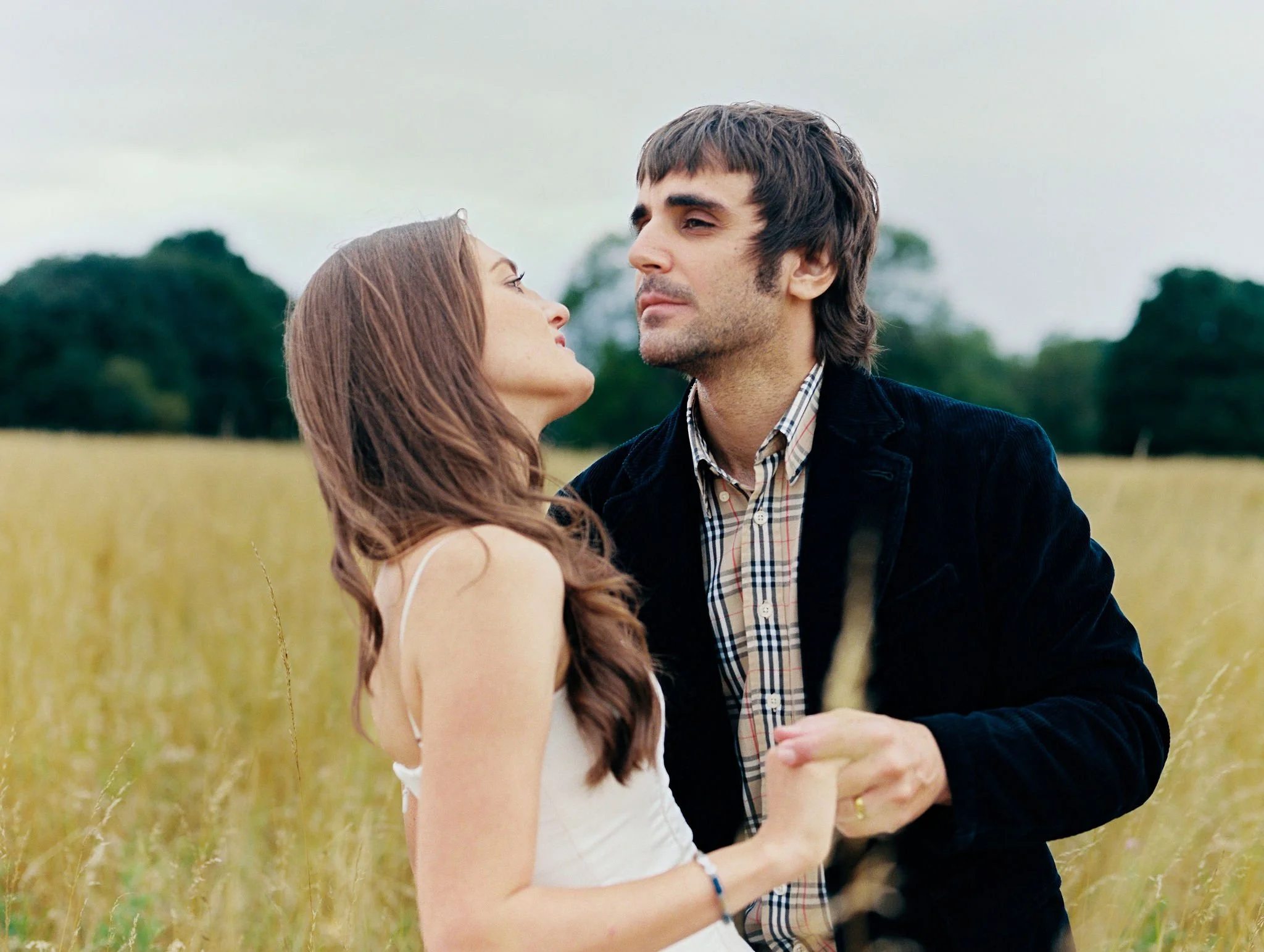A young woman and a young man are standing close together in a field with tall grass and trees in the background. The woman is looking up at the man, and they are holding hands.