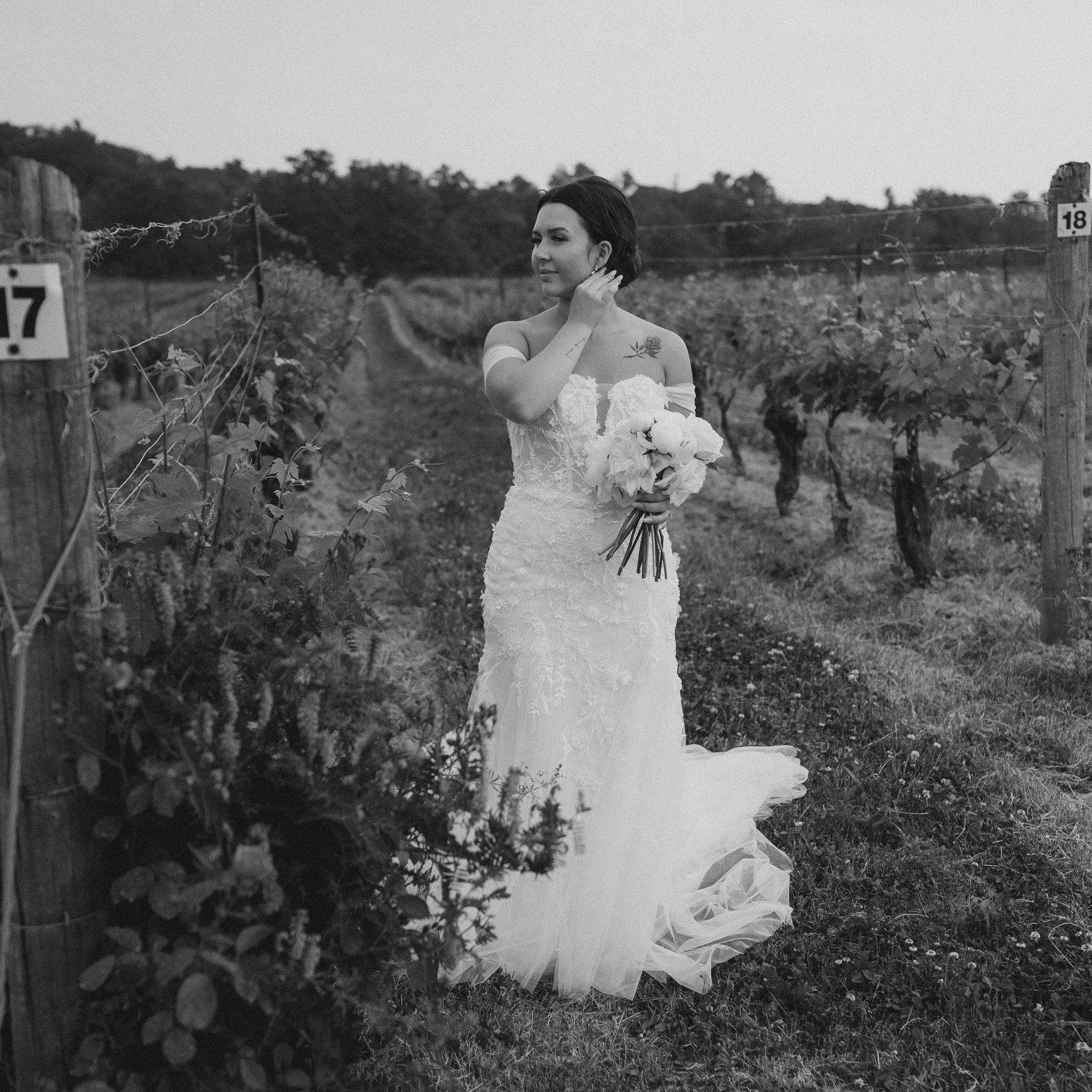 A woman in a wedding dress standing in a vineyard holding a bouquet of flowers.
