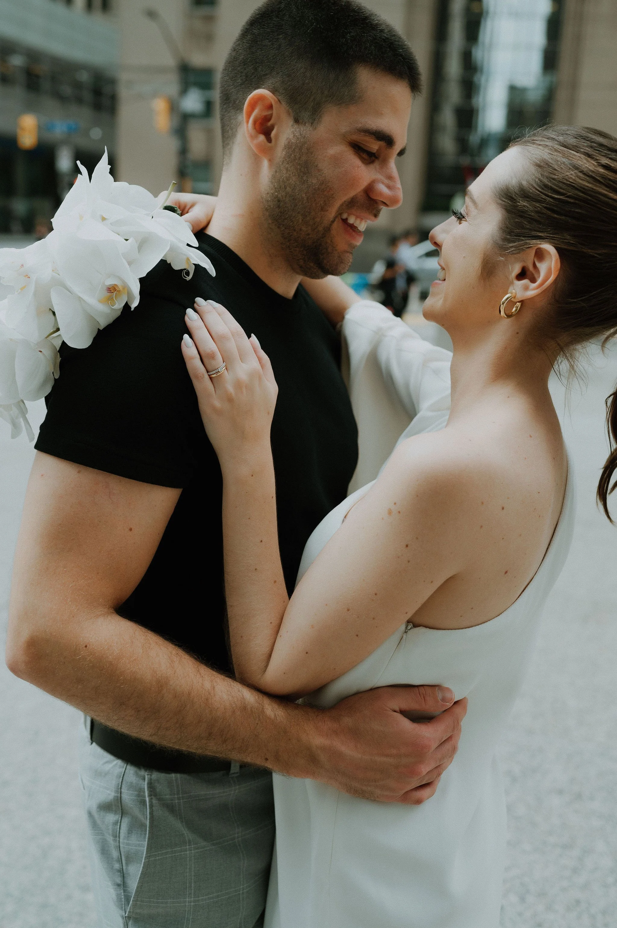 A couple is smiling and embracing each other on a city street, with the woman wearing a white dress and the man in a black t-shirt. The woman has her hands on the man's shoulders, showing an engagement ring, and the man is holding the woman around he