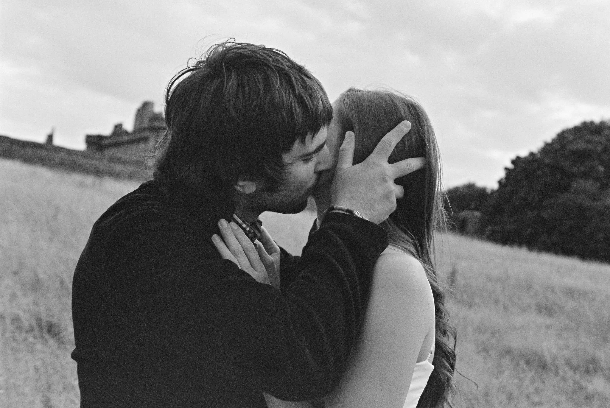A black and white photo of a man and woman kissing outdoors in a field, with trees and a building in the background.