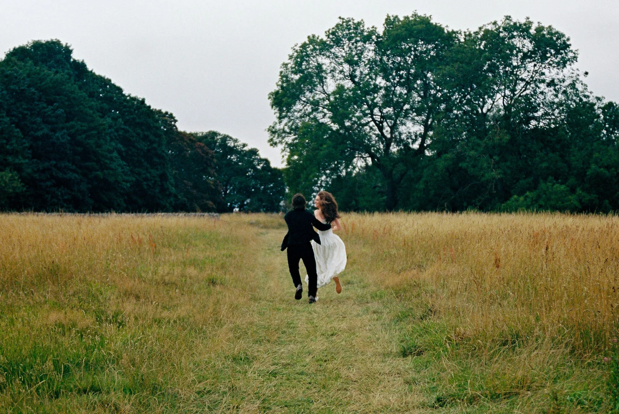 A couple dressed in formal attire, the woman in a white dress and the man in a black suit, running through a grassy field with trees in the background.