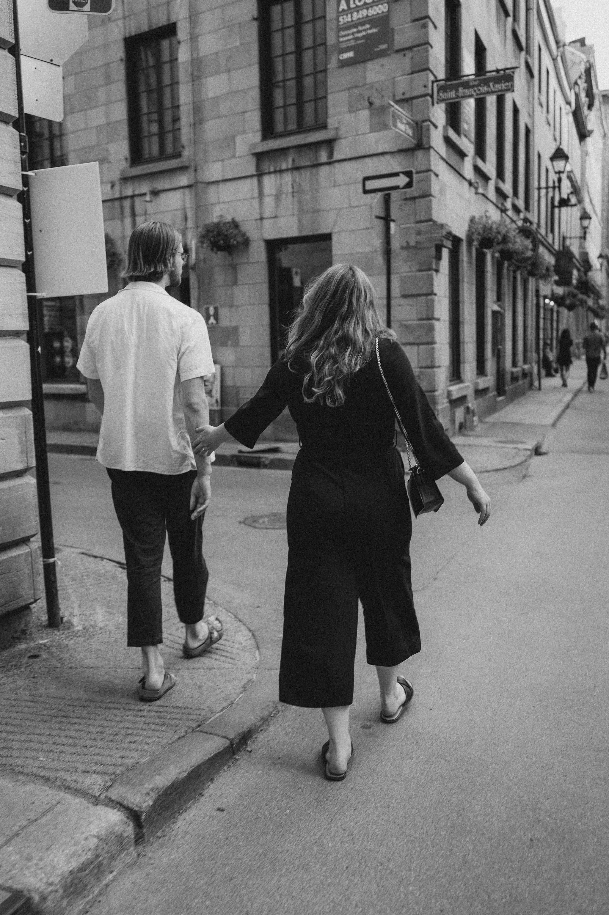 A black-and-white photo of a man and woman holding hands while crossing a city street. The man is wearing a light shirt and dark pants, while the woman is wearing a dark top and cropped pants. They are walking past old buildings with flower baskets a