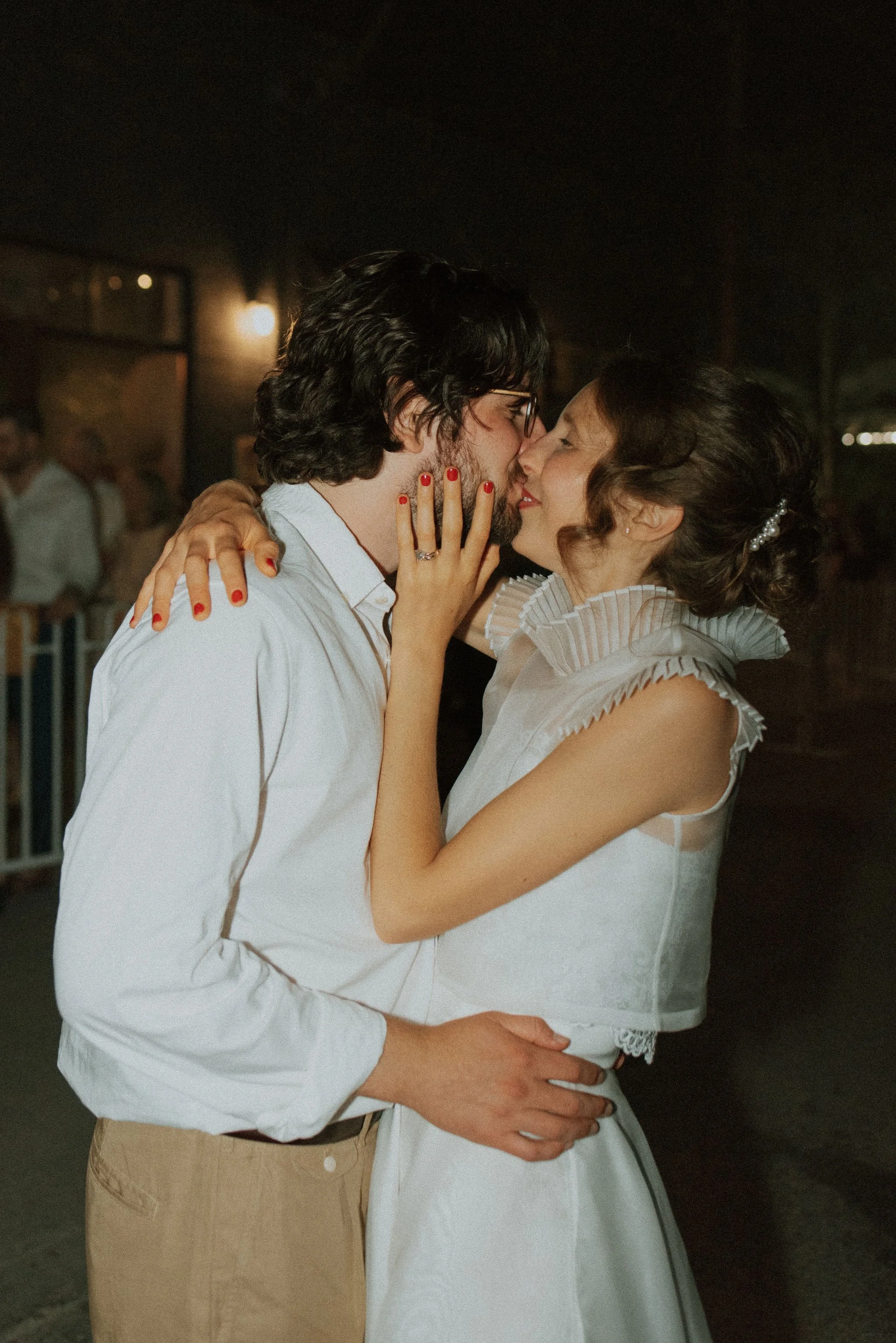 A couple shares a kiss at nighttime, with the woman wearing a white dress and the man wearing a white shirt and khakis, embracing each other lovingly.