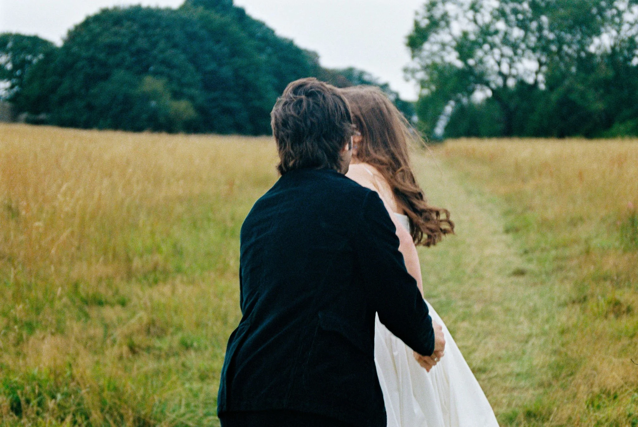 A couple standing in a grassy field, embracing each other, with trees in the background.