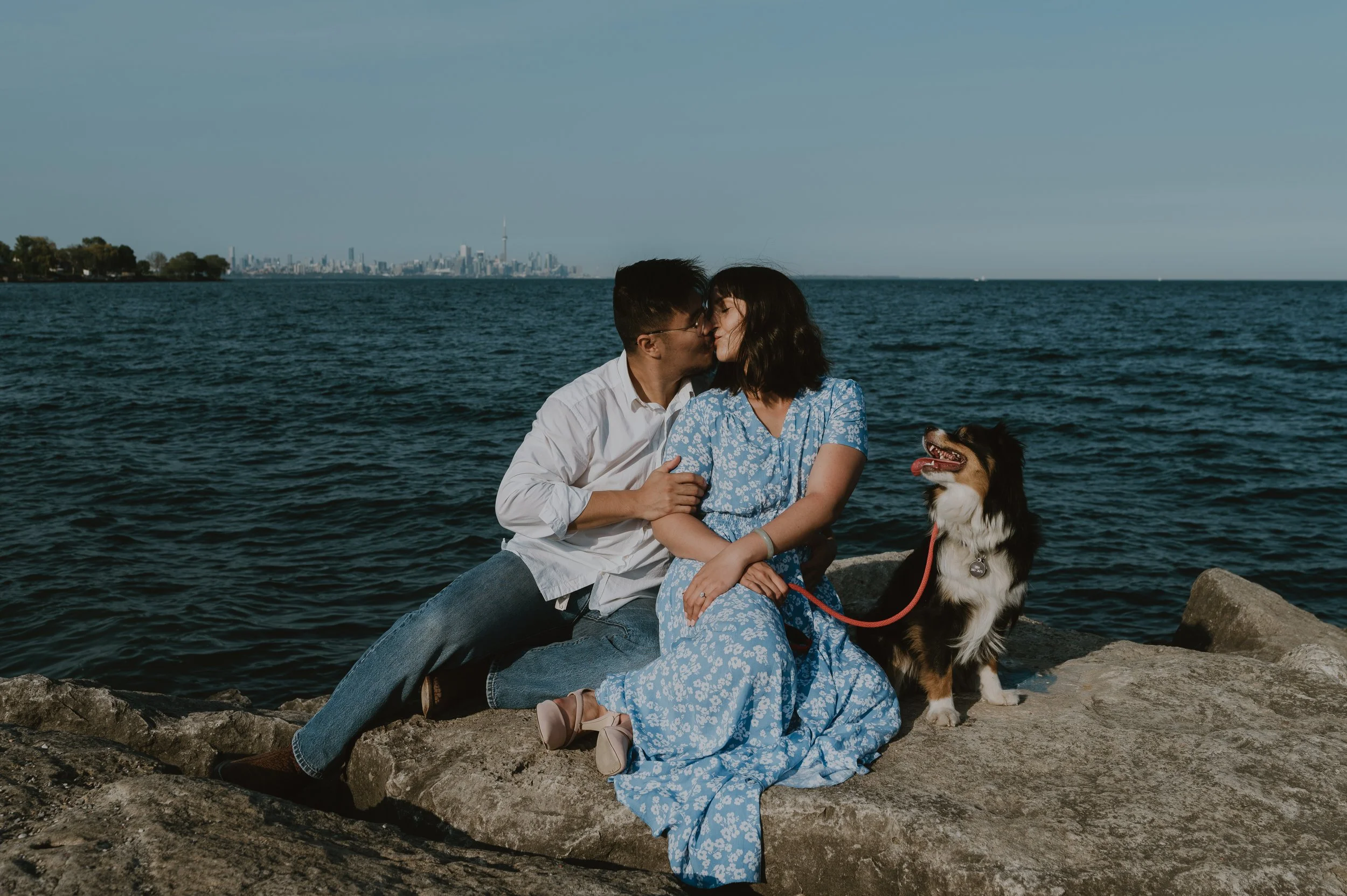 A couple sharing a kiss while sitting on a rock by the water with their dog, with the city skyline in the background.