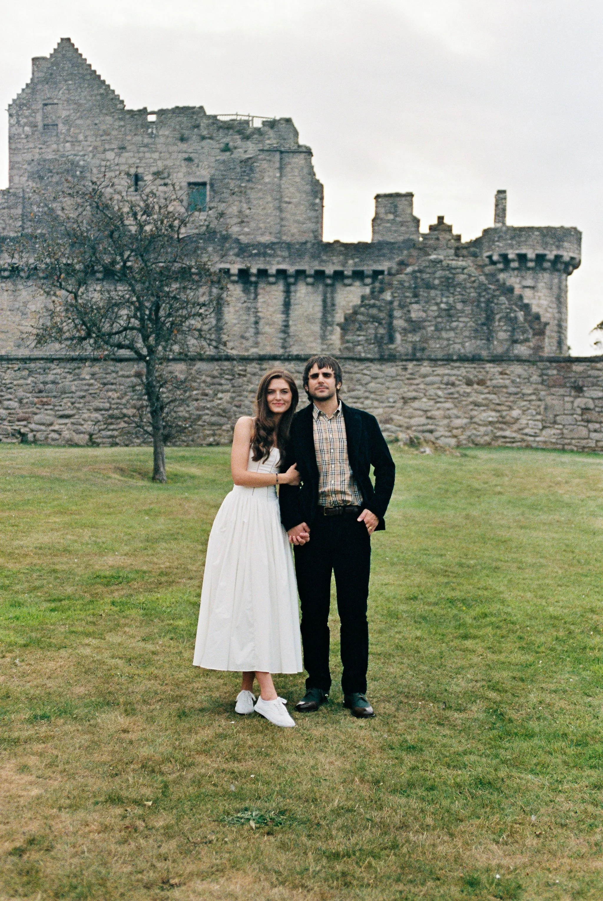 A young couple standing arm-in-arm on a grassy lawn in front of an old stone castle during daytime. The woman is wearing a white dress and sneakers, while the man is dressed in dark pants, a blazer, and a plaid shirt.
