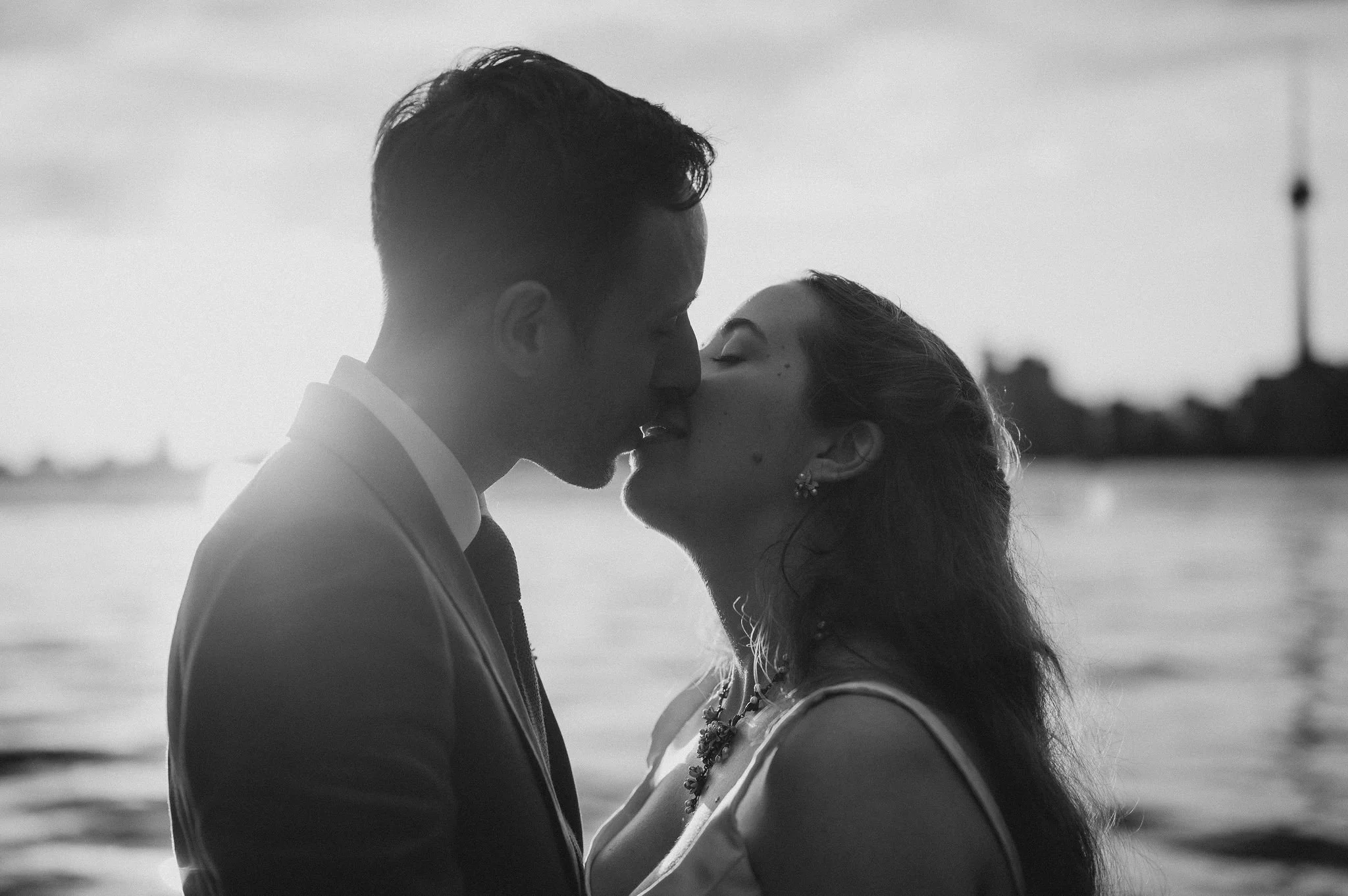 A black-and-white photograph of a couple kissing by the waterfront during sunset, with city skyline and water in the background.