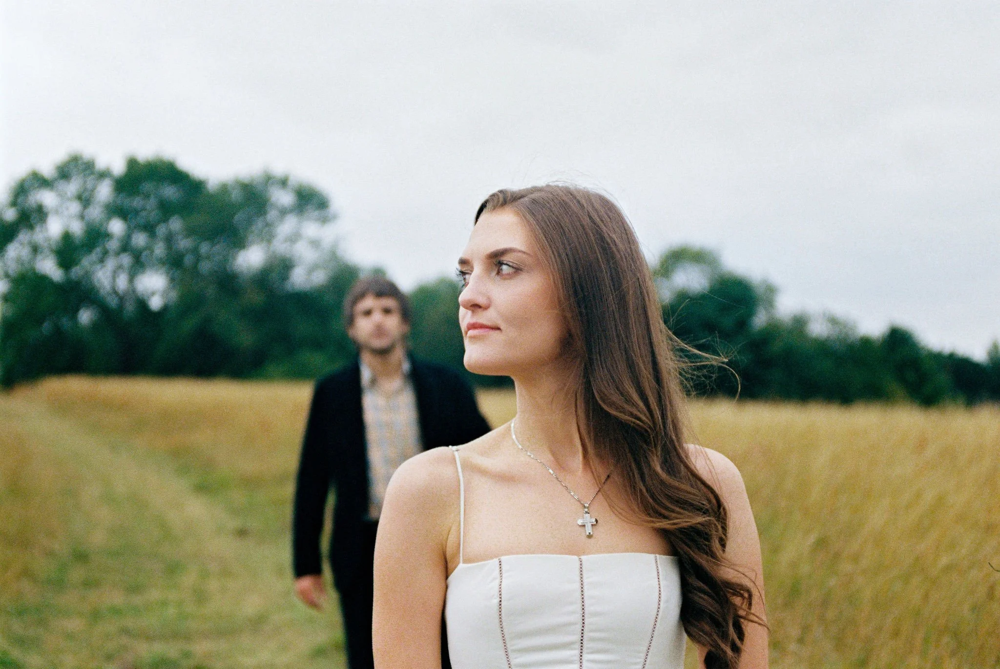 A woman with long brown hair in a white dress with spaghetti straps, wearing a cross necklace, standing in a field with tall grass, with a man in a dark jacket and checkered shirt in the background, across from her.