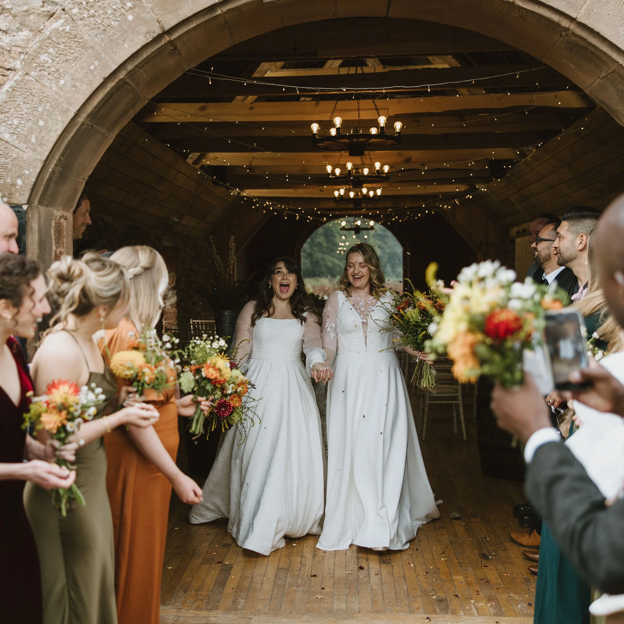 Two women in wedding dresses holding hands and smiling, surrounded by friends and family holding bouquets, inside a rustic venue with string lights and wooden ceiling.