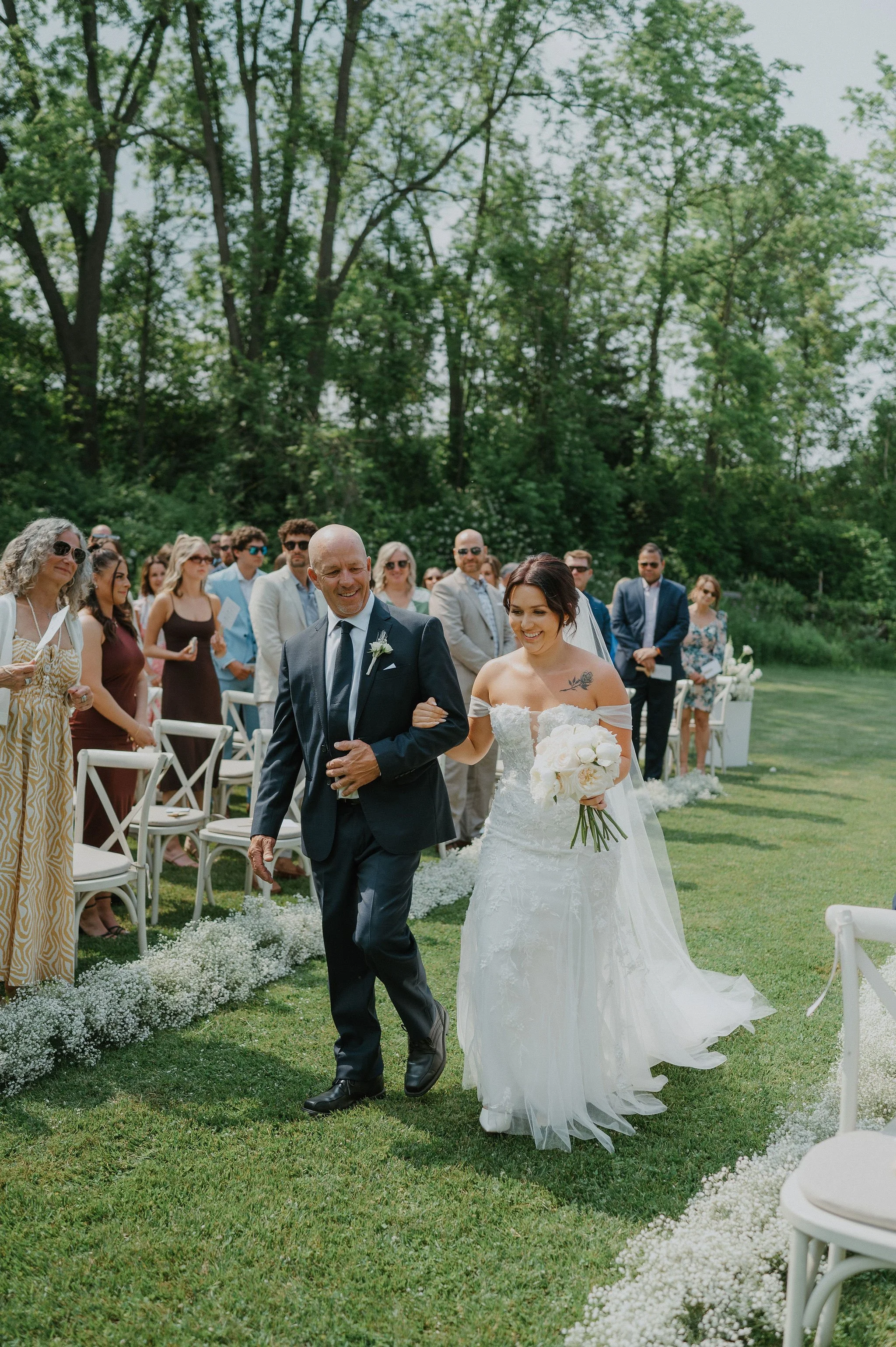 A bride walks down the aisle with her father at an outdoor wedding, surrounded by seated guests and lush green trees.