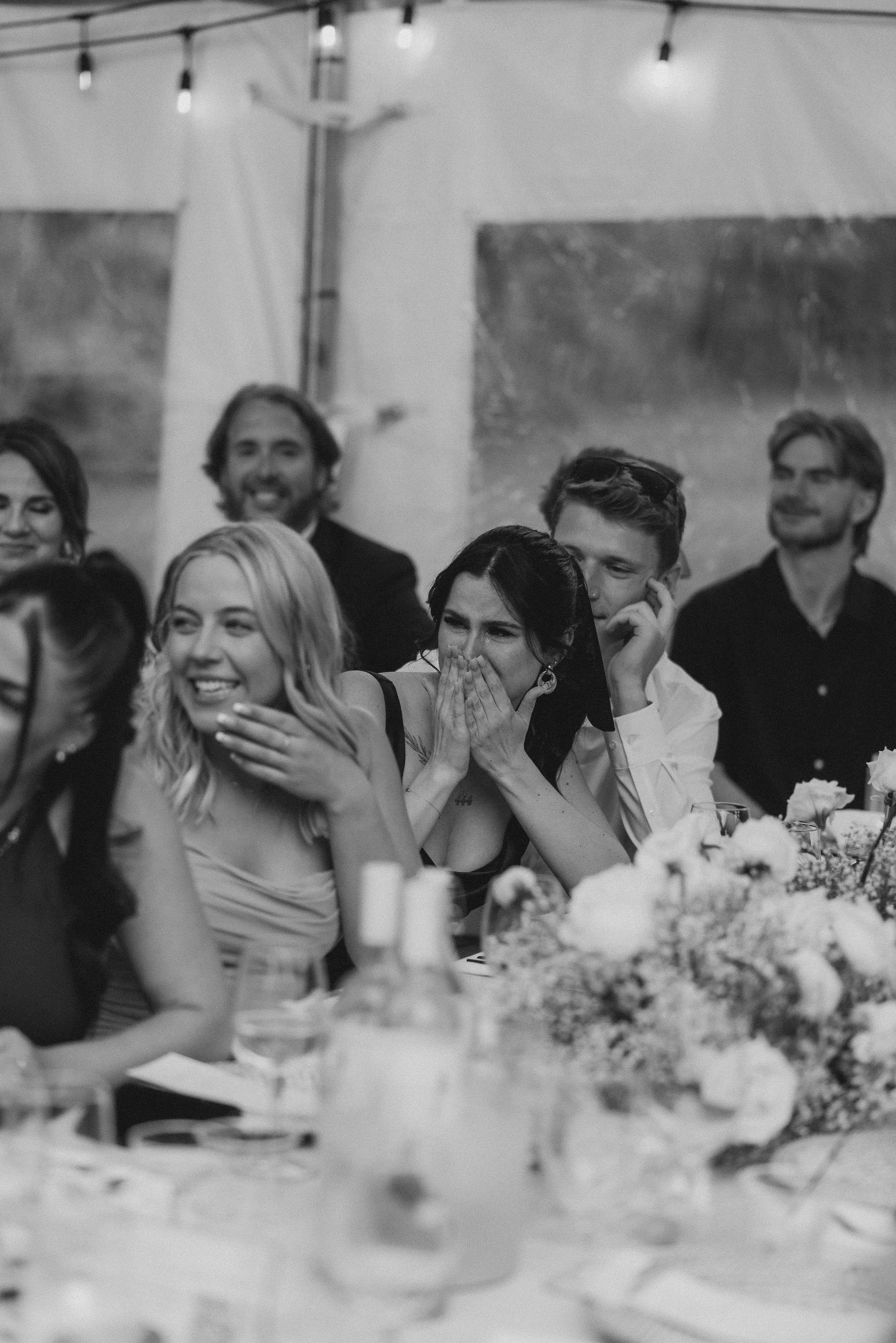 A group of people seated at a table, with some people smiling and one woman covering her mouth in surprise, at a celebration or gathering indoors.