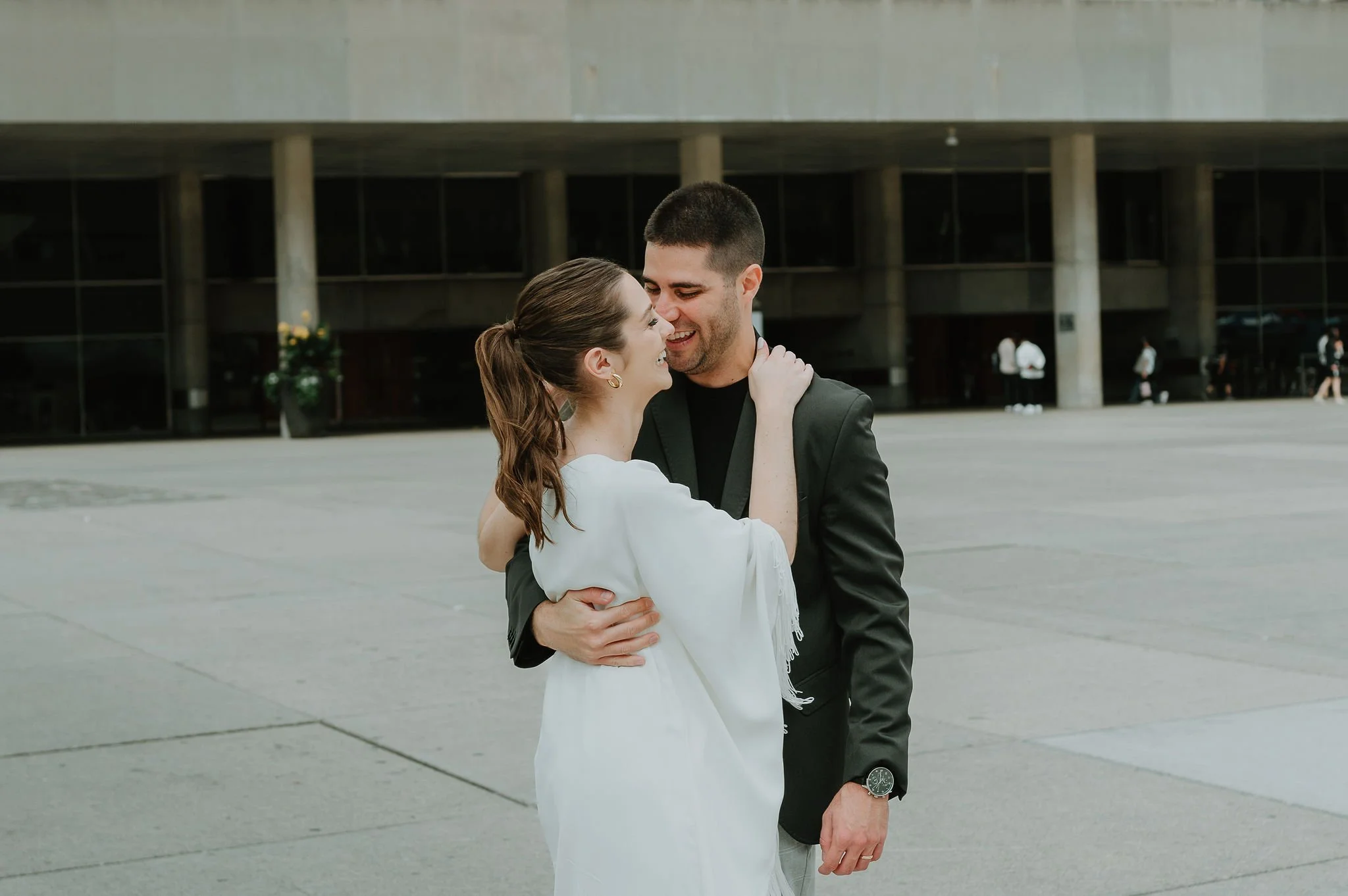 A couple is embracing and smiling at each other outdoors in front of a modern building. The woman has brown hair in a ponytail and is wearing a white dress, while the man has short dark hair, a beard, and is dressed in a black blazer and shirt.