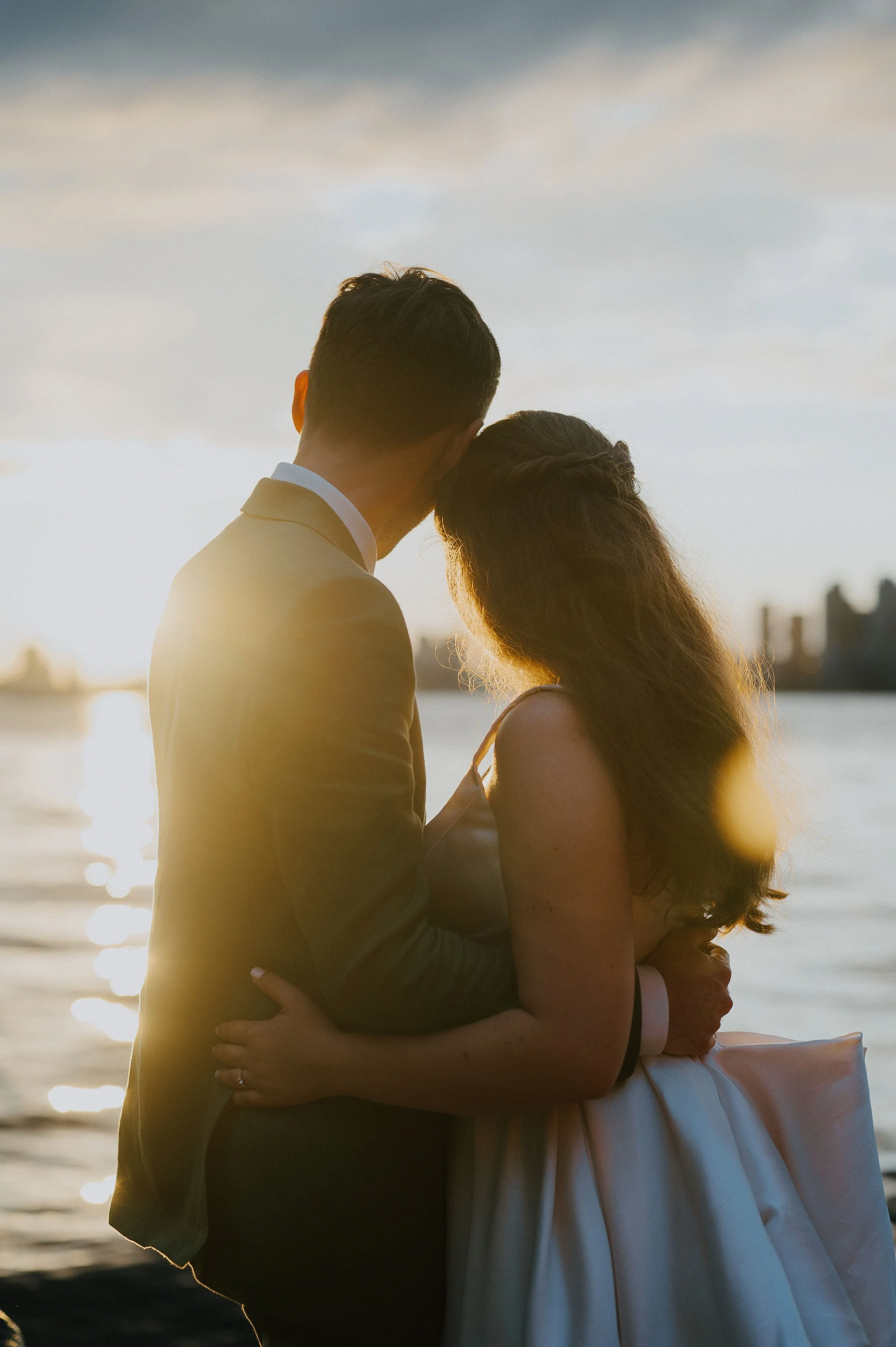 A couple dressed in wedding attire, embracing by a river at sunset, with city skyline in the background.