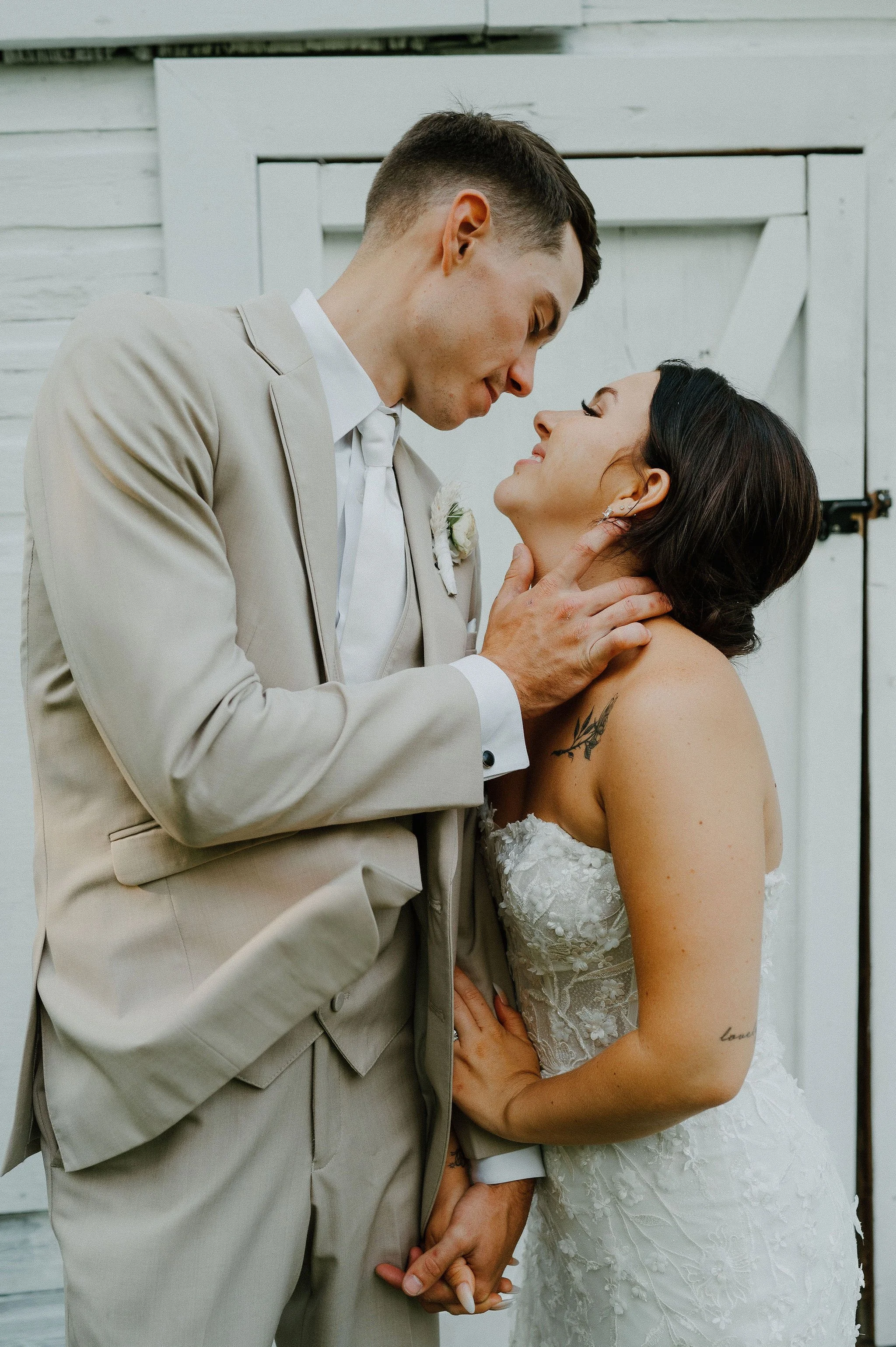 A bride and groom share a kiss at their wedding, with the groom holding the bride's chin and the bride looking up at him, both smiling gently.
