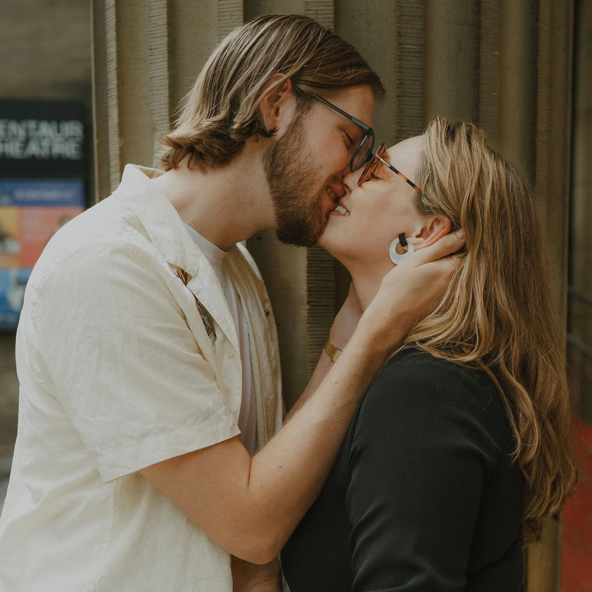 A couple sharing a kiss on a city street with a wall in the background.