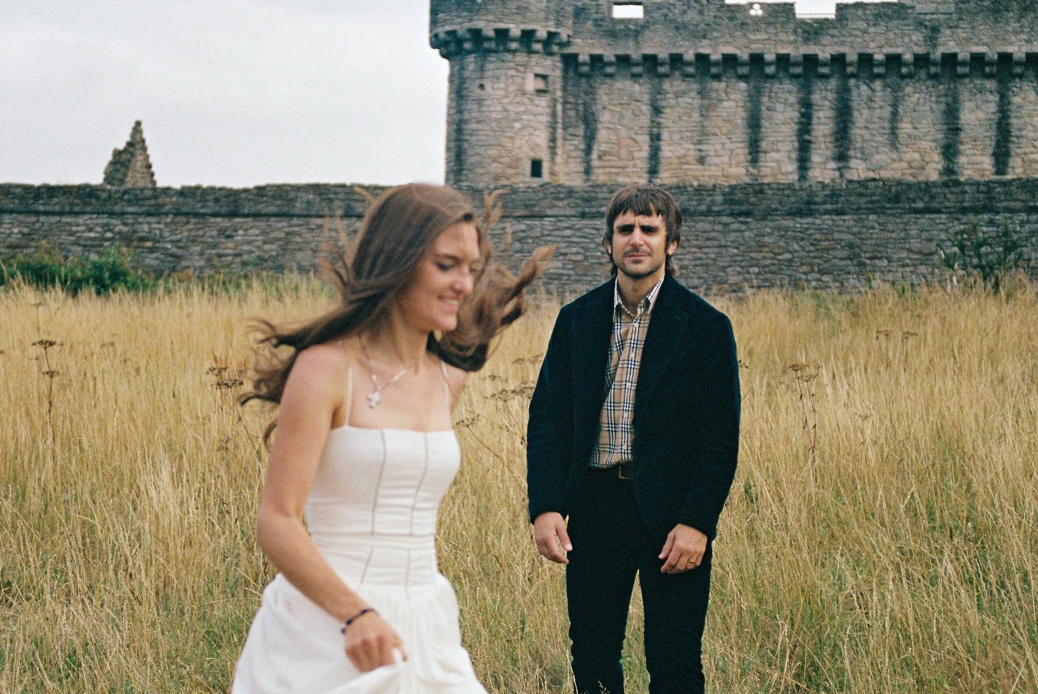 A woman in a white dress walking in a grassy field with a man in a black jacket and plaid shirt standing in the background near an old stone castle.