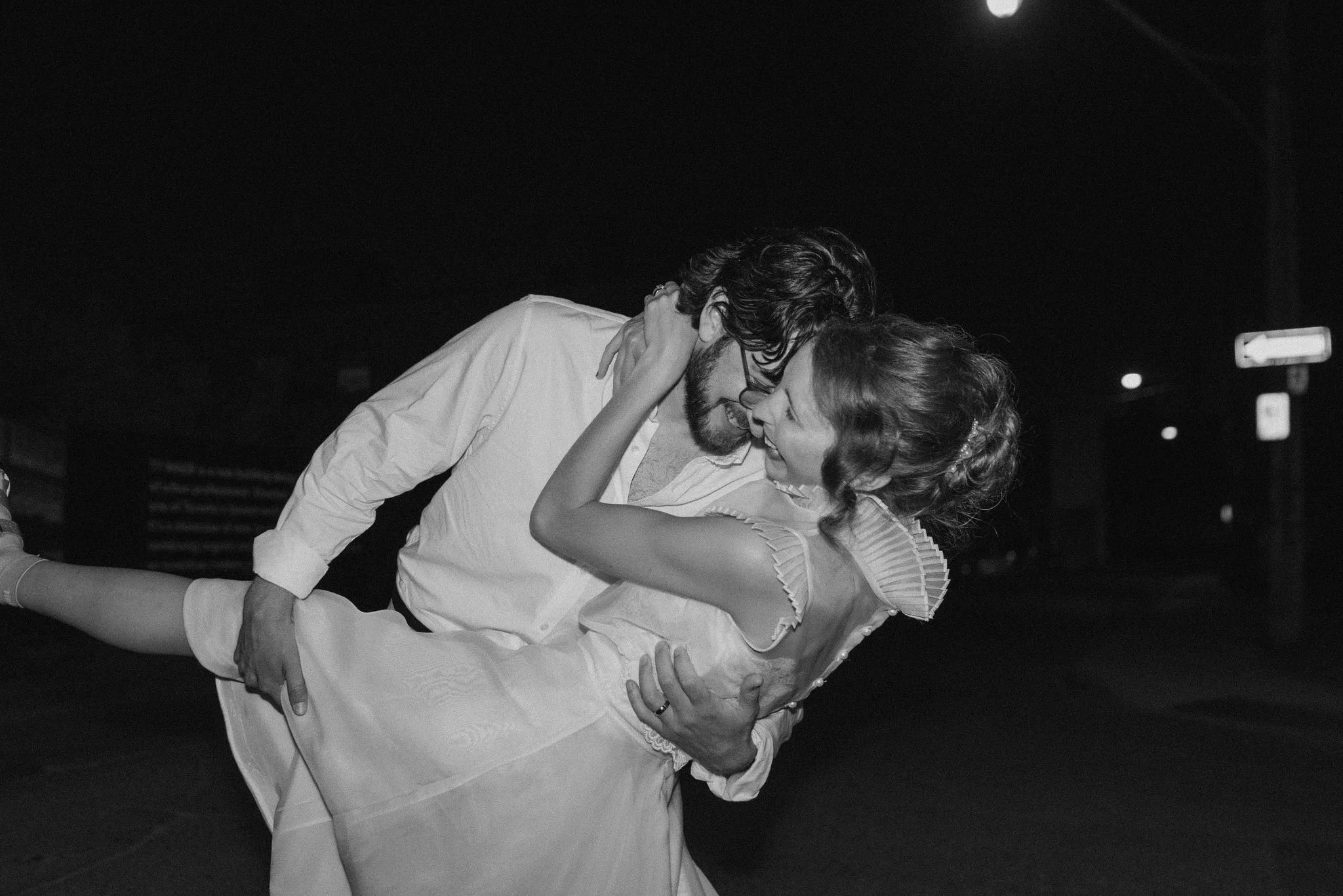 A man in a white shirt and a woman in a dress are dancing closely together at night outside, with the man lifting the woman who is smiling and looking at him.