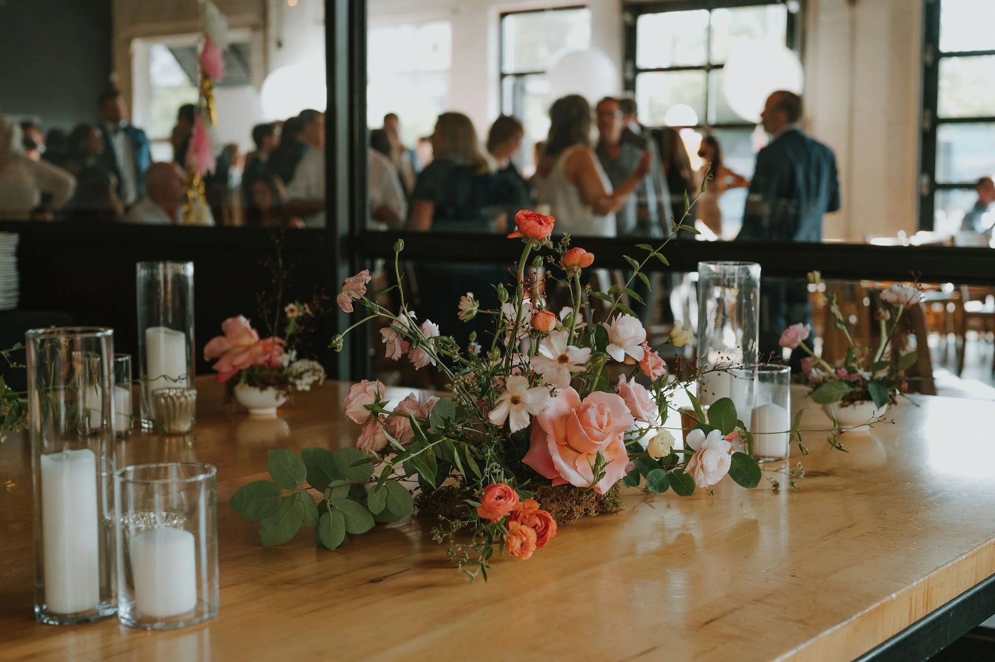 A floral centerpiece with pink and white flowers and green leaves on a wooden table, surrounded by white candles in glass holders, at an indoor event with people in the background.