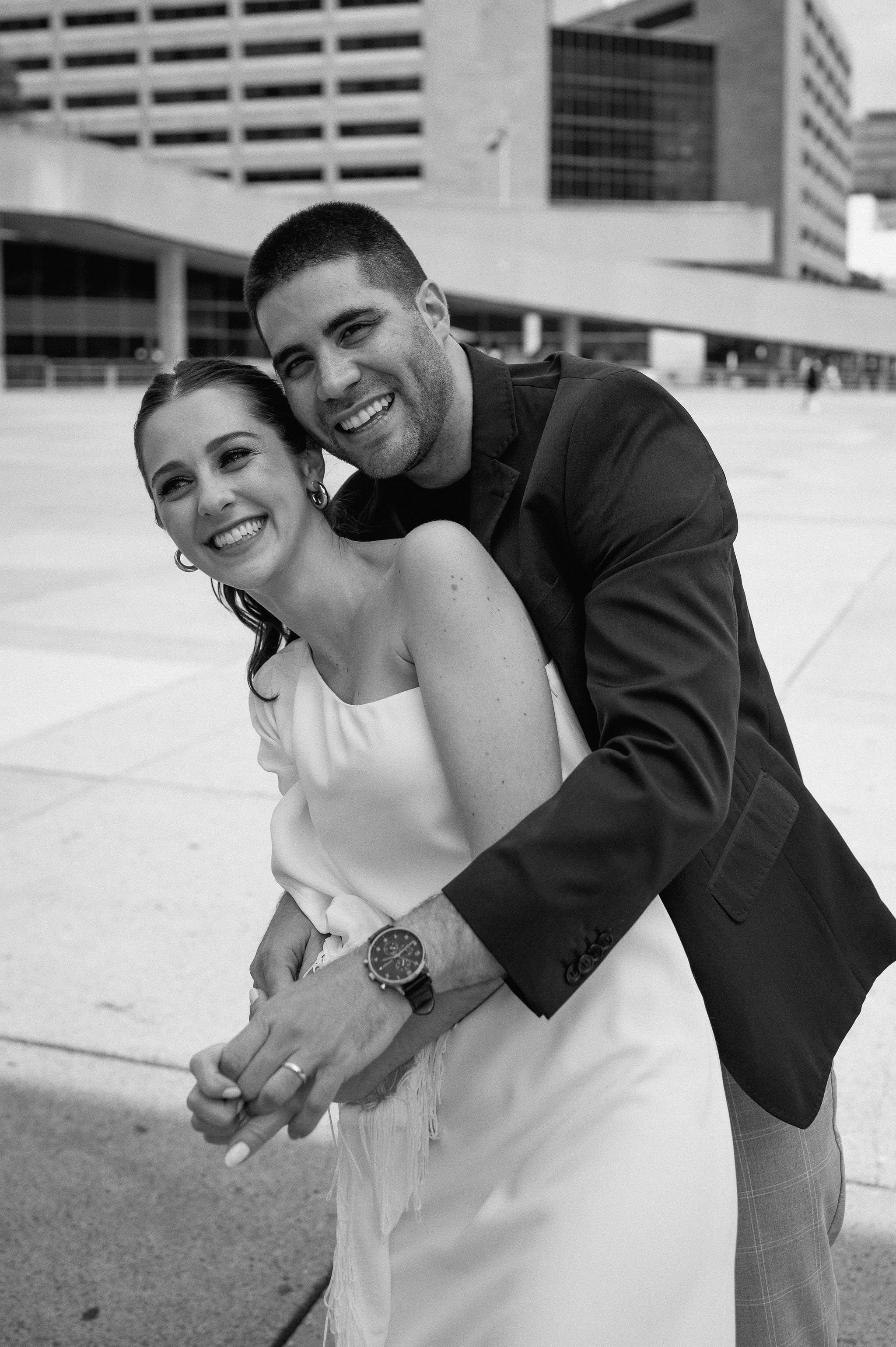 A happy couple in formal attire smiling and embracing in an outdoor urban setting, with large modern buildings in the background.