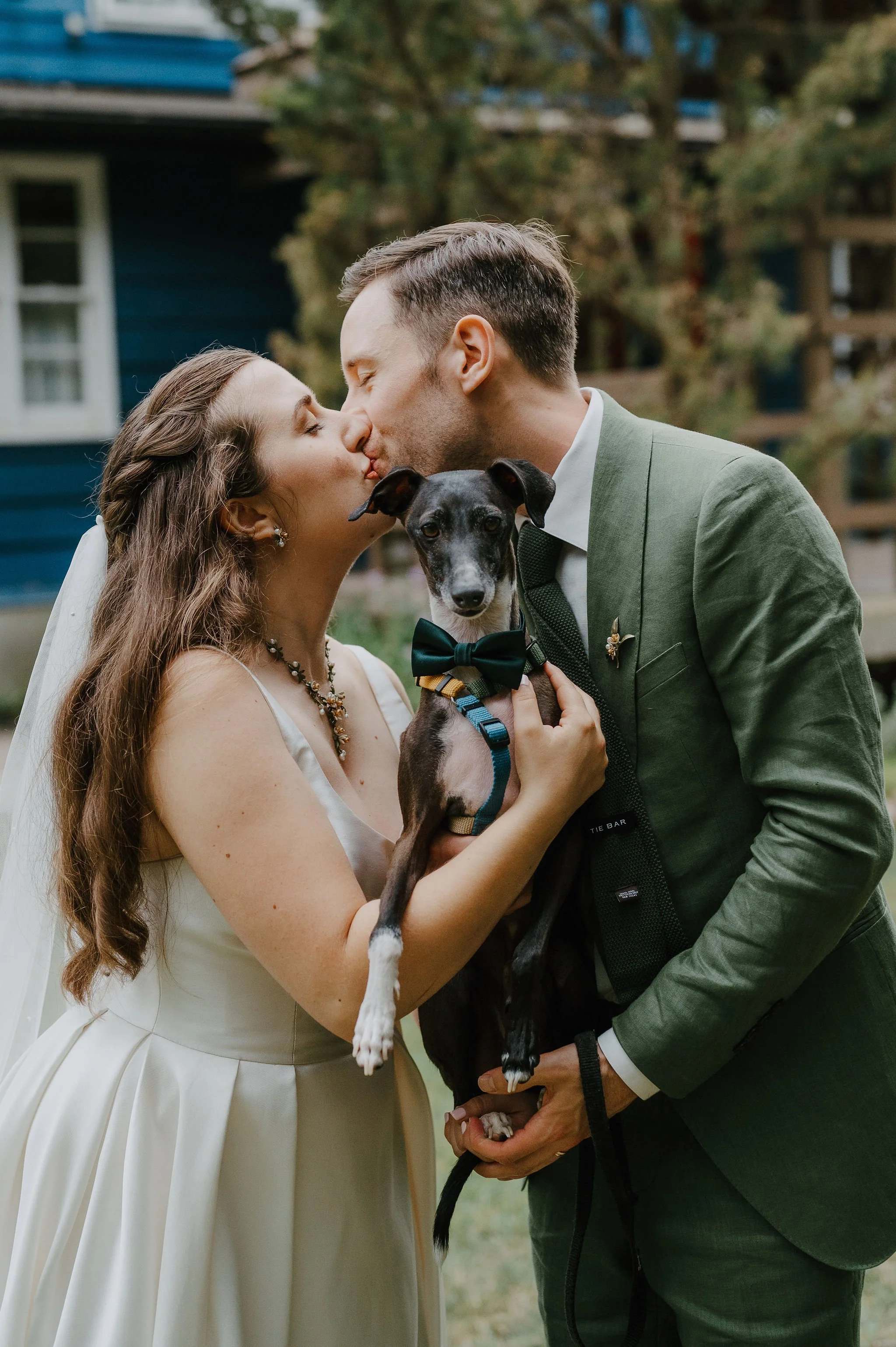 A newlywed couple kissing, holding their dog dressed with a bow tie, outdoors in front of a blue house and trees.
