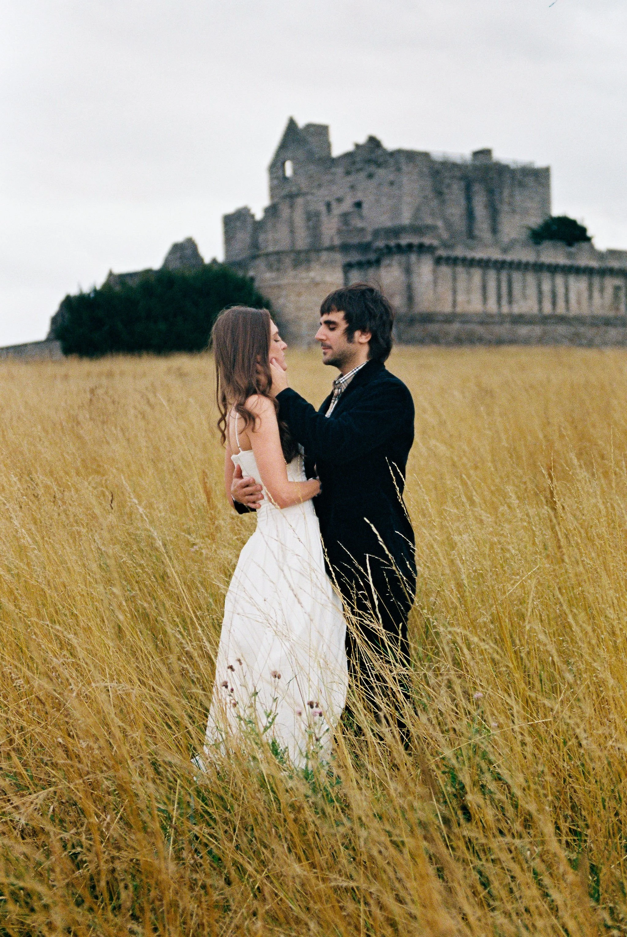 A couple stands in a field of tall grass with an ancient castle in the background.