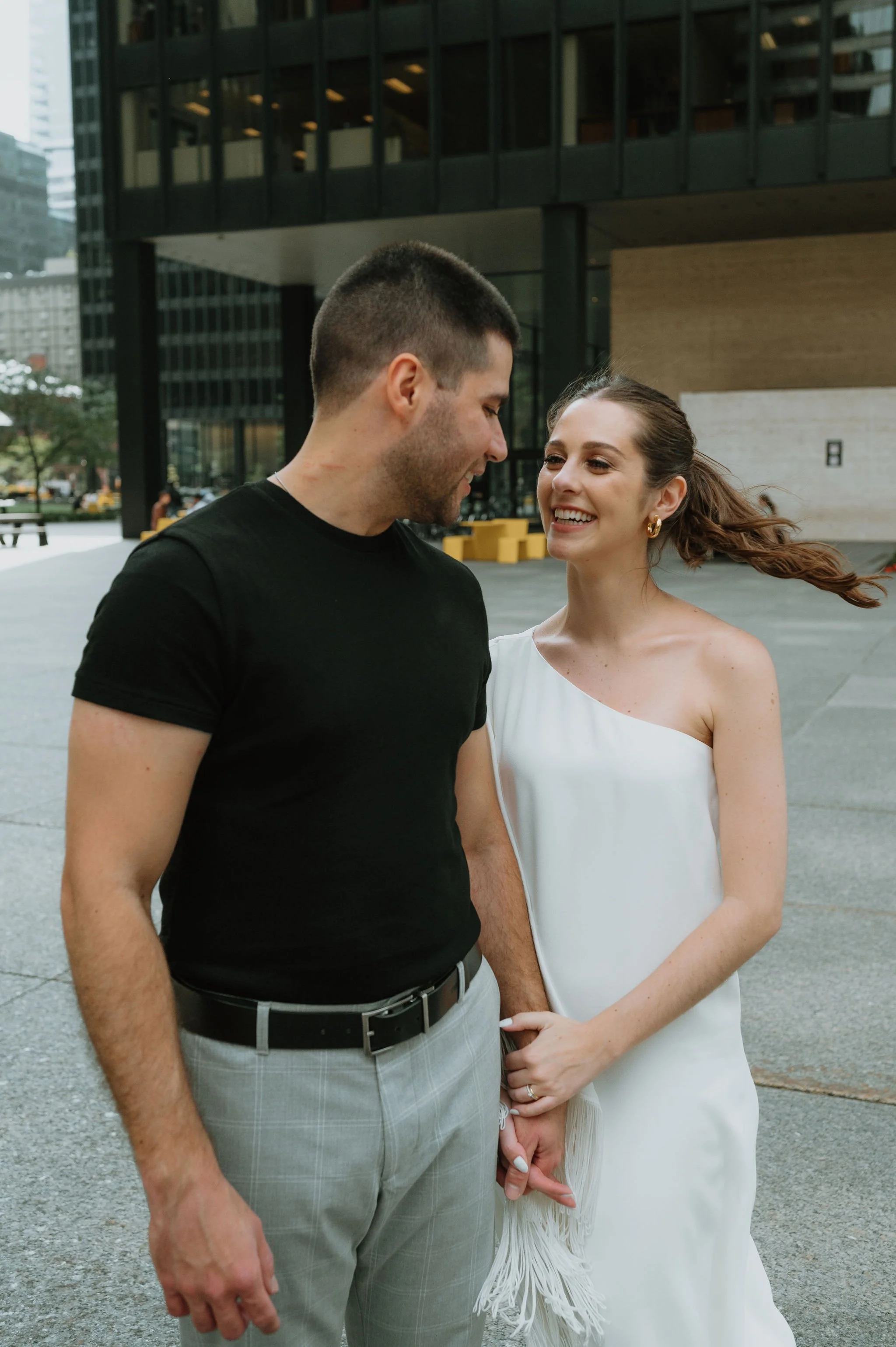 A smiling woman in a white dress holding hands with a man in a black t-shirt and gray pants, outside in an urban setting with modern buildings.