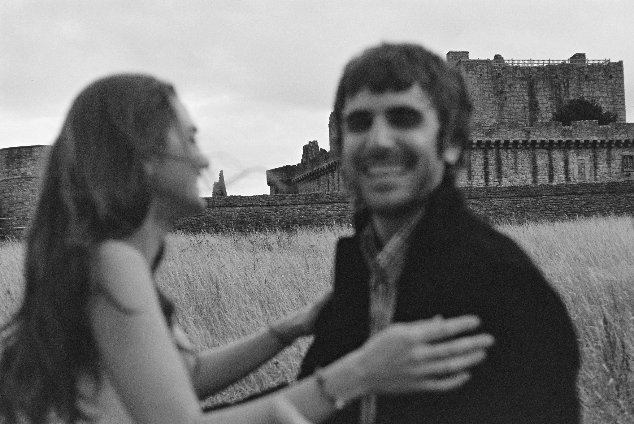 A blurry black and white photo of a couple outdoors in front of a castle or fortress, with the woman reaching out toward the man who is smiling and wearing sunglasses.