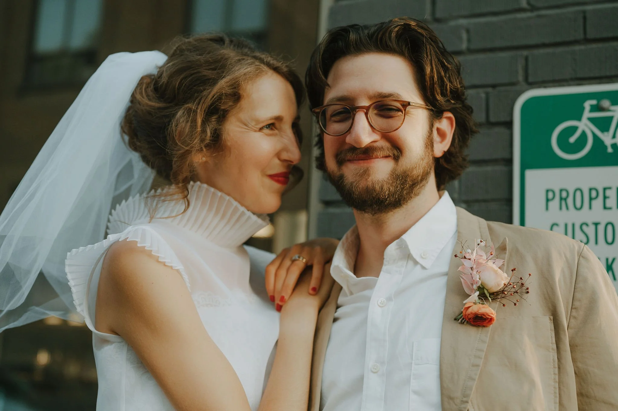 A bride and groom smiling together outdoors, with the bride wearing a white dress and veil and the groom in a beige suit with a boutonniere.
