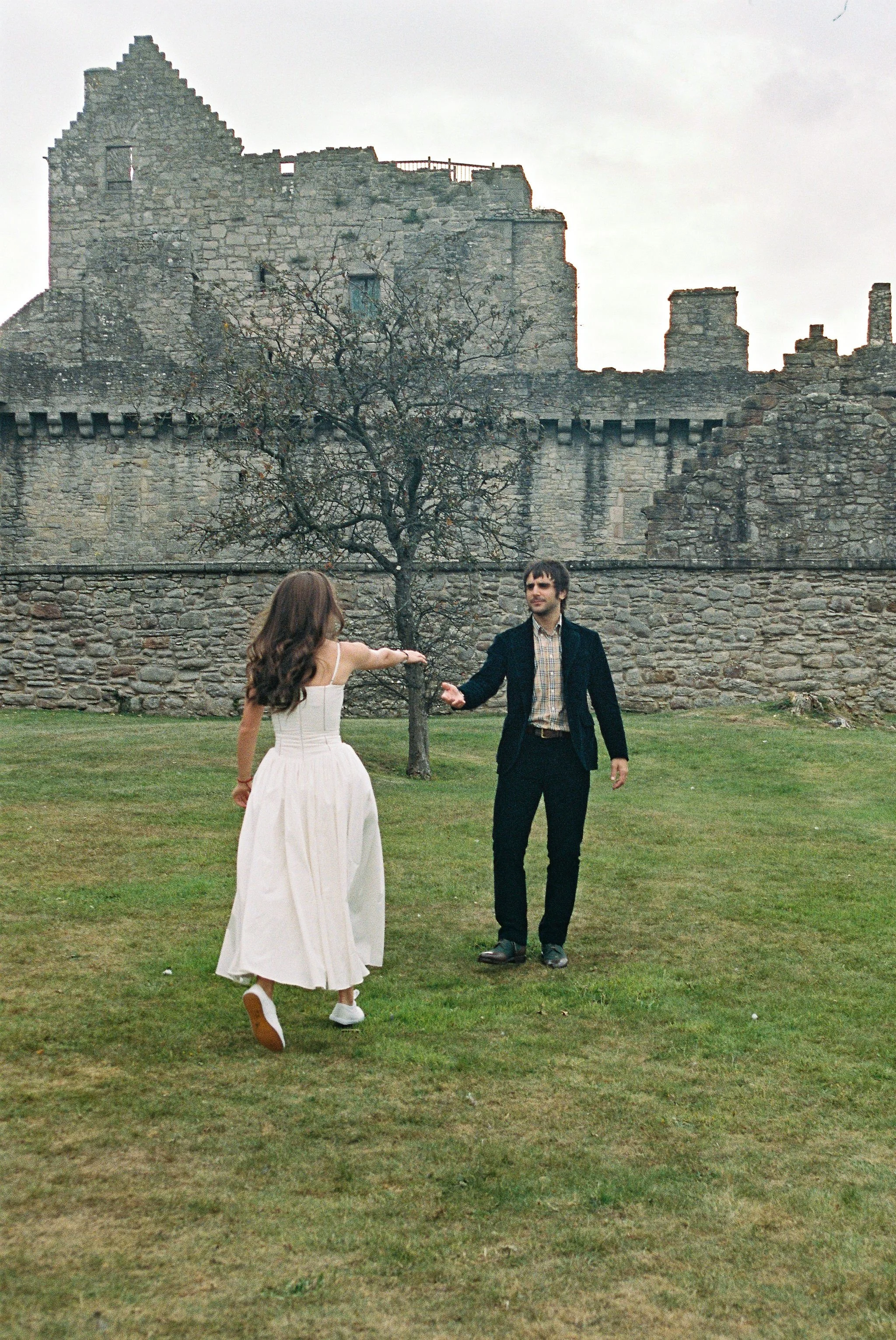 A woman in a white dress appears to be dancing or twirling with a man wearing a black blazer and plaid shirt, standing on a grassy area in front of an old stone castle wall with a leafless tree.
