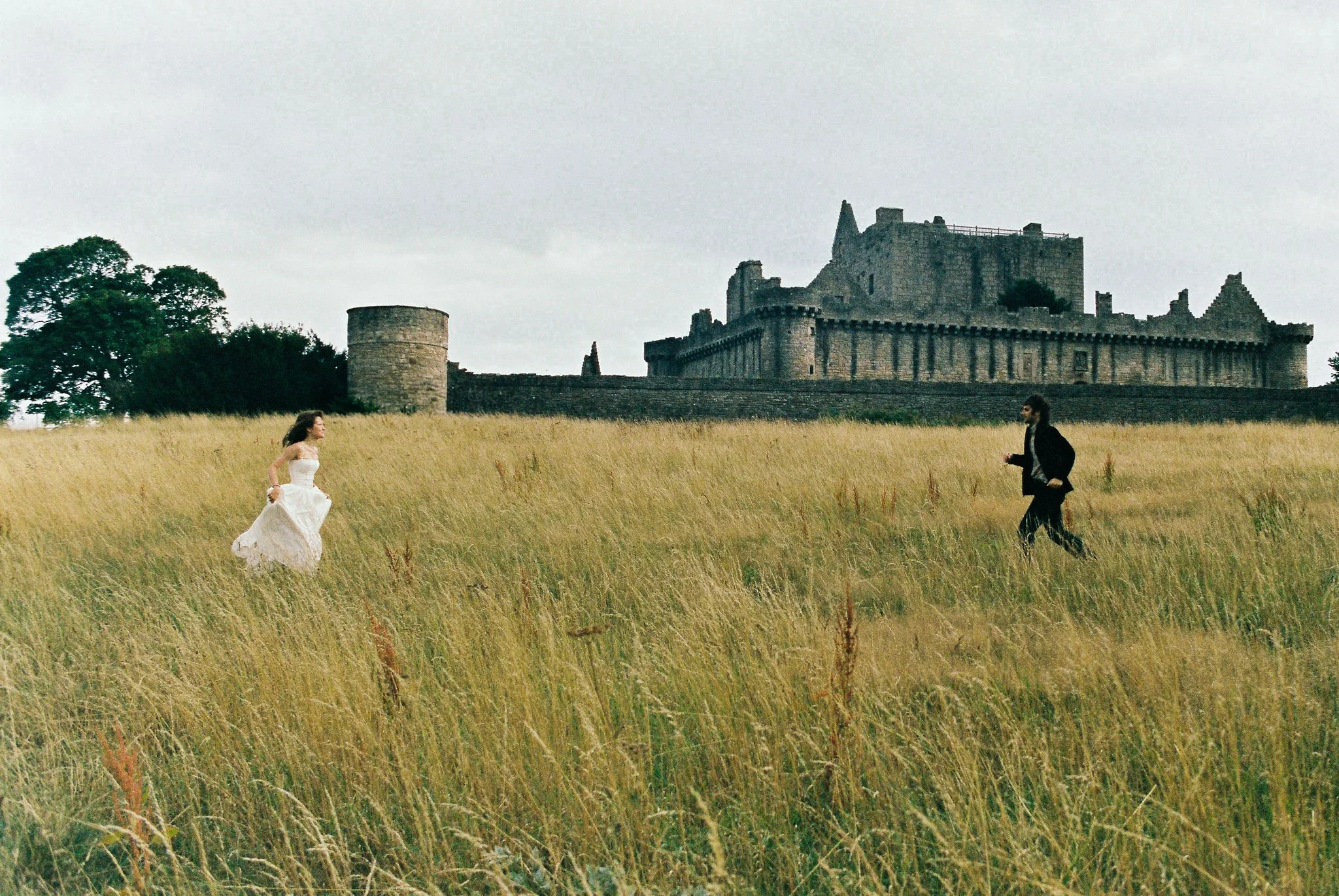 A woman in a white dress running towards a man in a black suit across a field of tall grass with a large historic castle in the background.